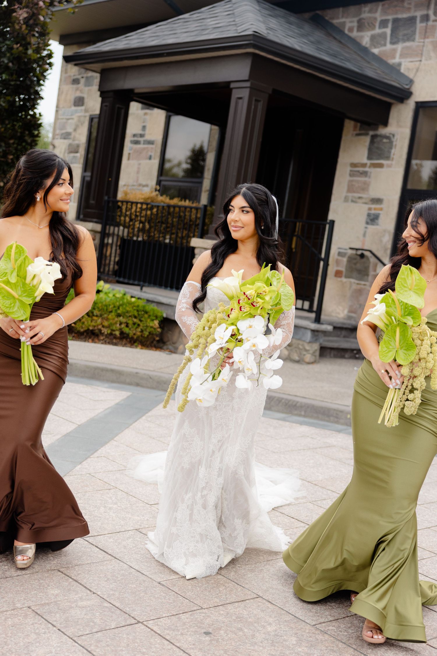 Wedding party members in brown and olive green dresses pose with white floral bouquets outside brick building.