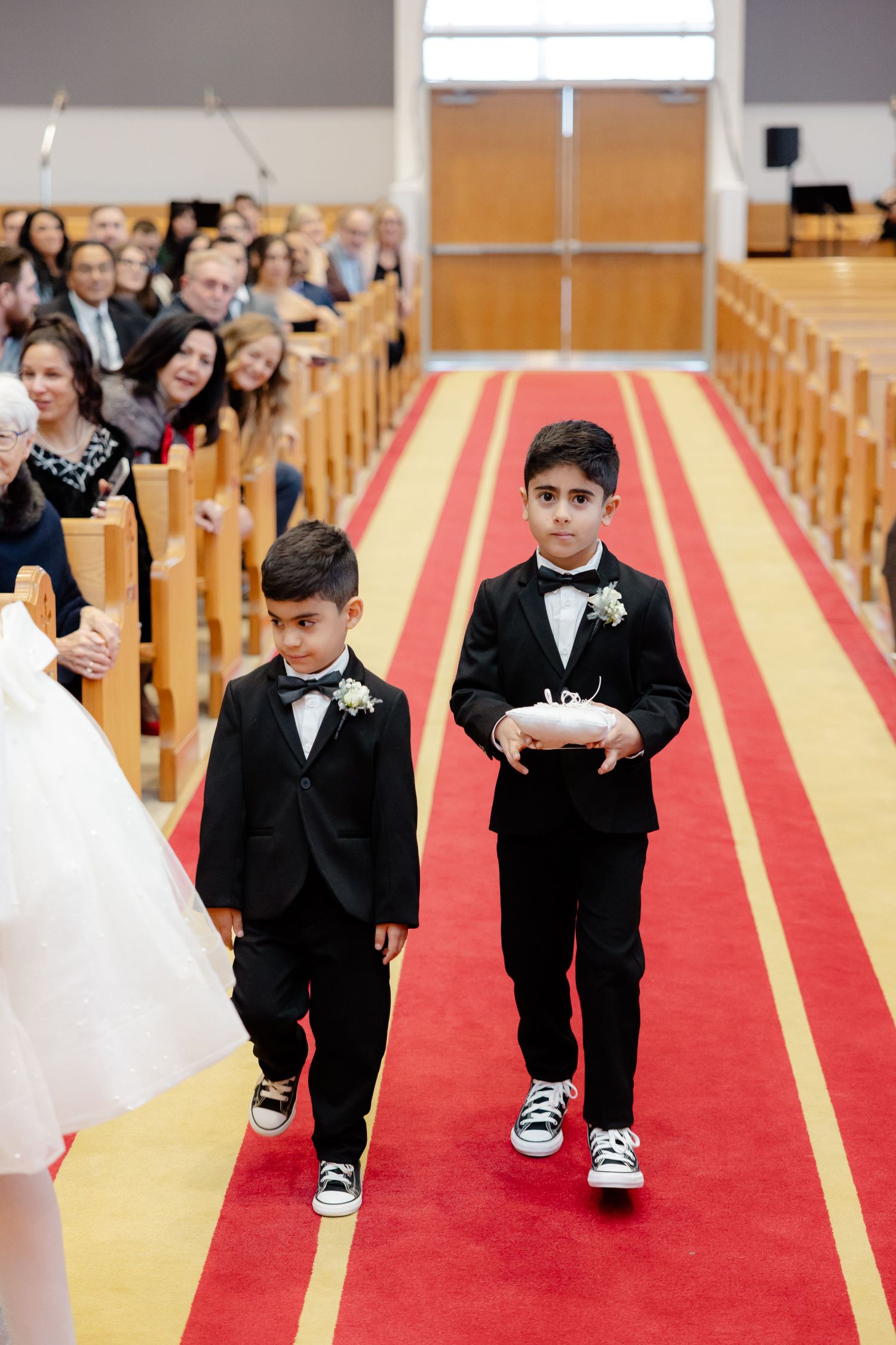 Two young attendants in black suits walk down the church aisle carrying rings.