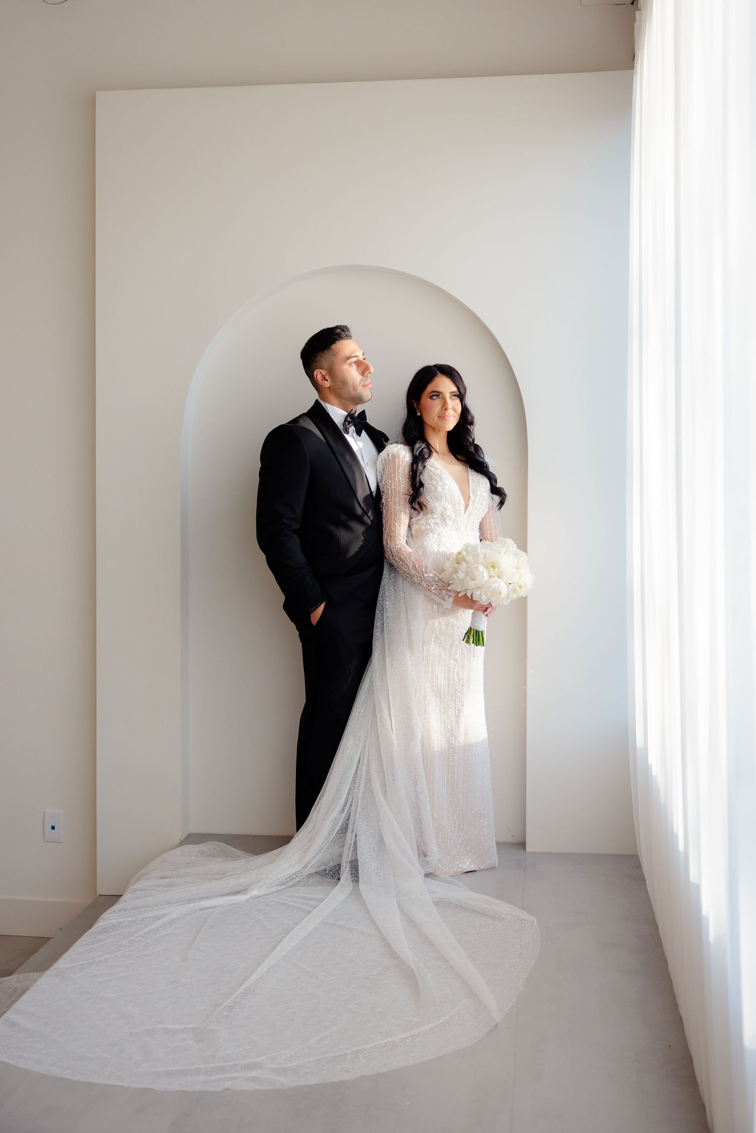 Wedding couple poses together in front of white archway, bride wearing lace gown with flowing veil.