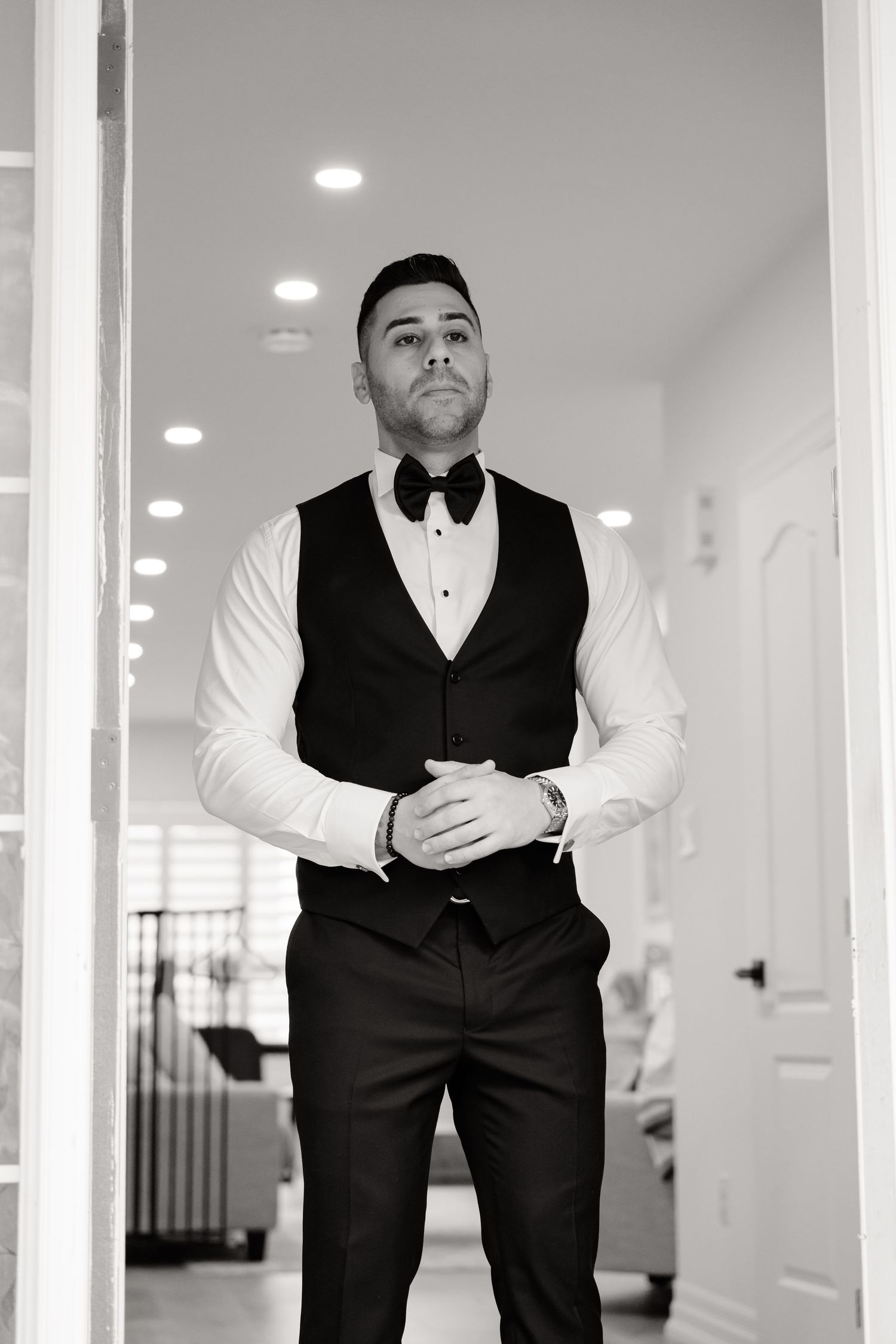 A formal black and white portrait of a groom in a tuxedo vest and bow tie standing in a hallway.