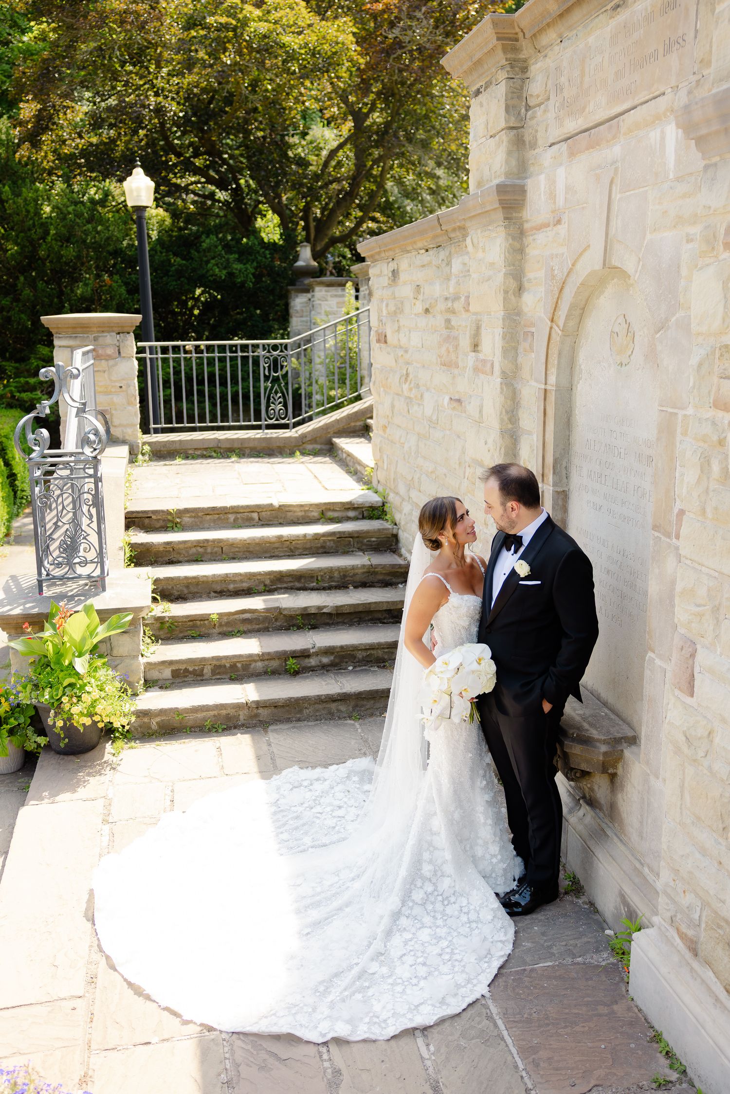 A romantic wedding couple embracing near stone steps and columns in an elegant garden setting.