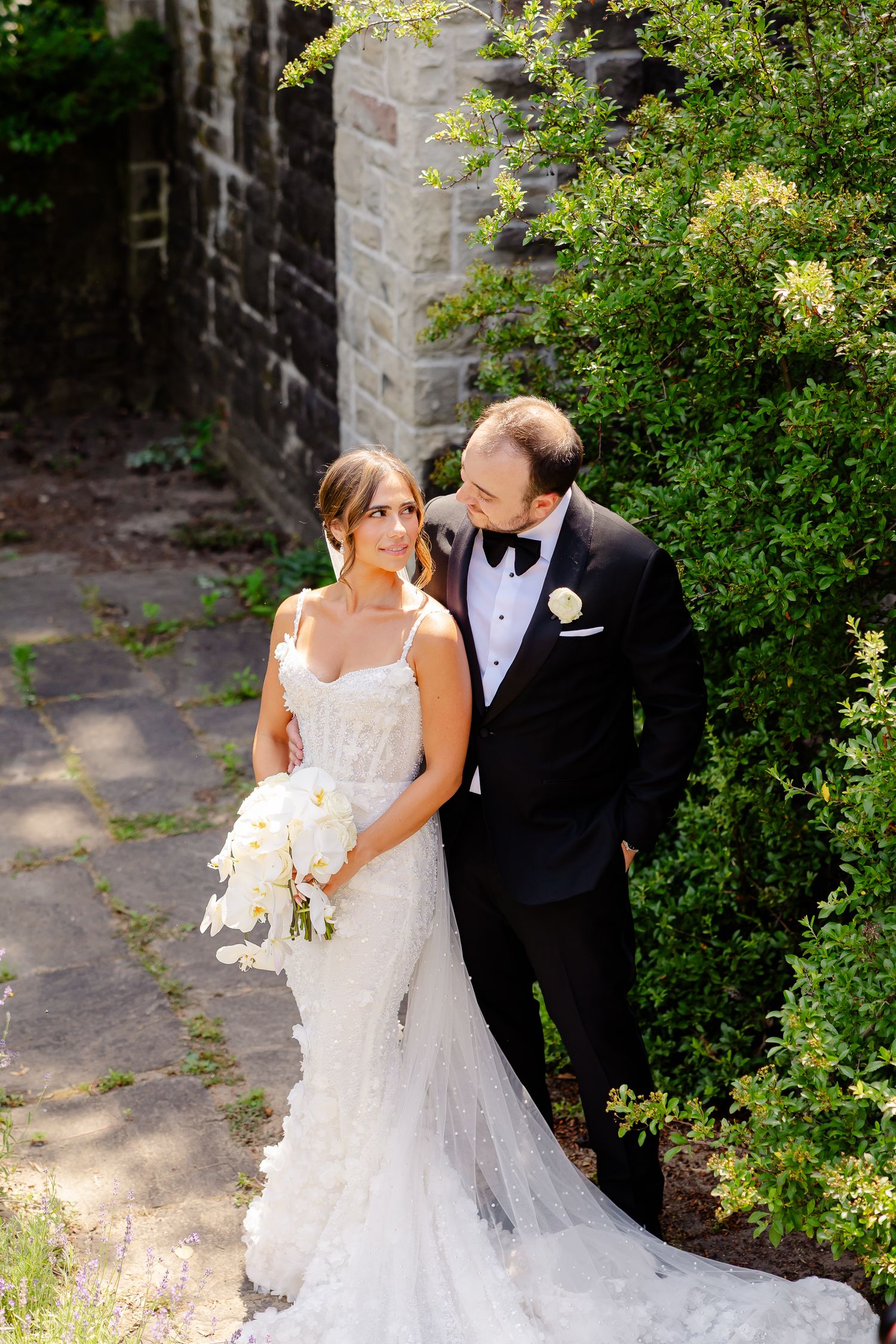 A couple in formal wedding attire stands together in a garden setting with stone walls and lush greenery.