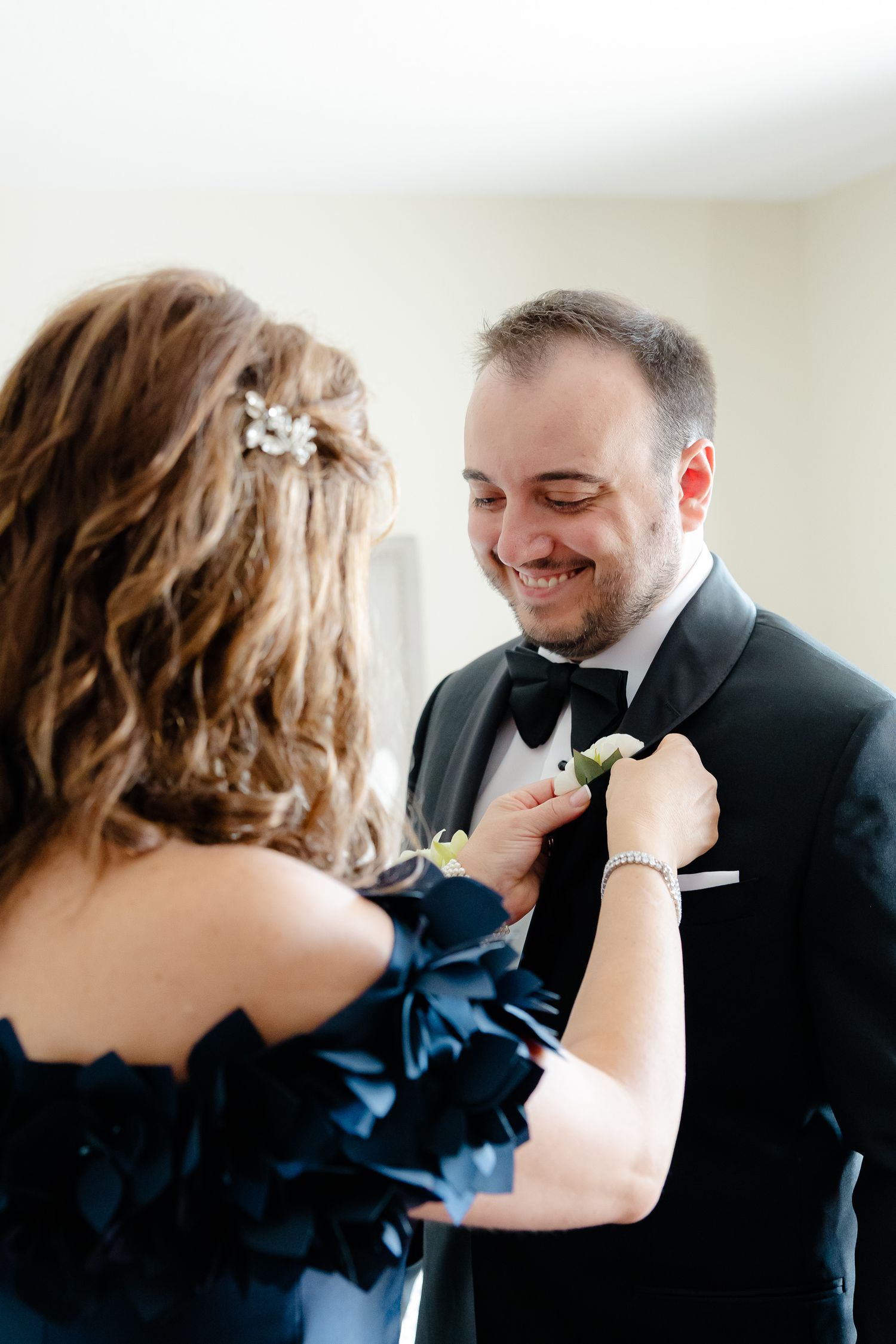 A person in navy blue dress helps adjust the boutonniere on a smiling groom's black tuxedo lapel.