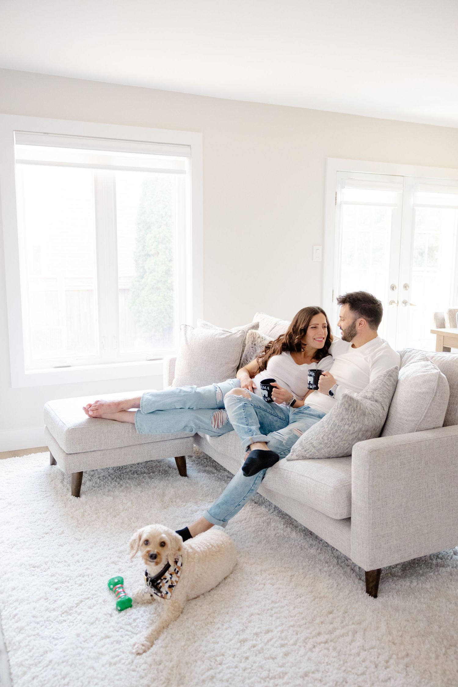 Couple relaxing on couch with dog, enjoying cozy living room with large windows.