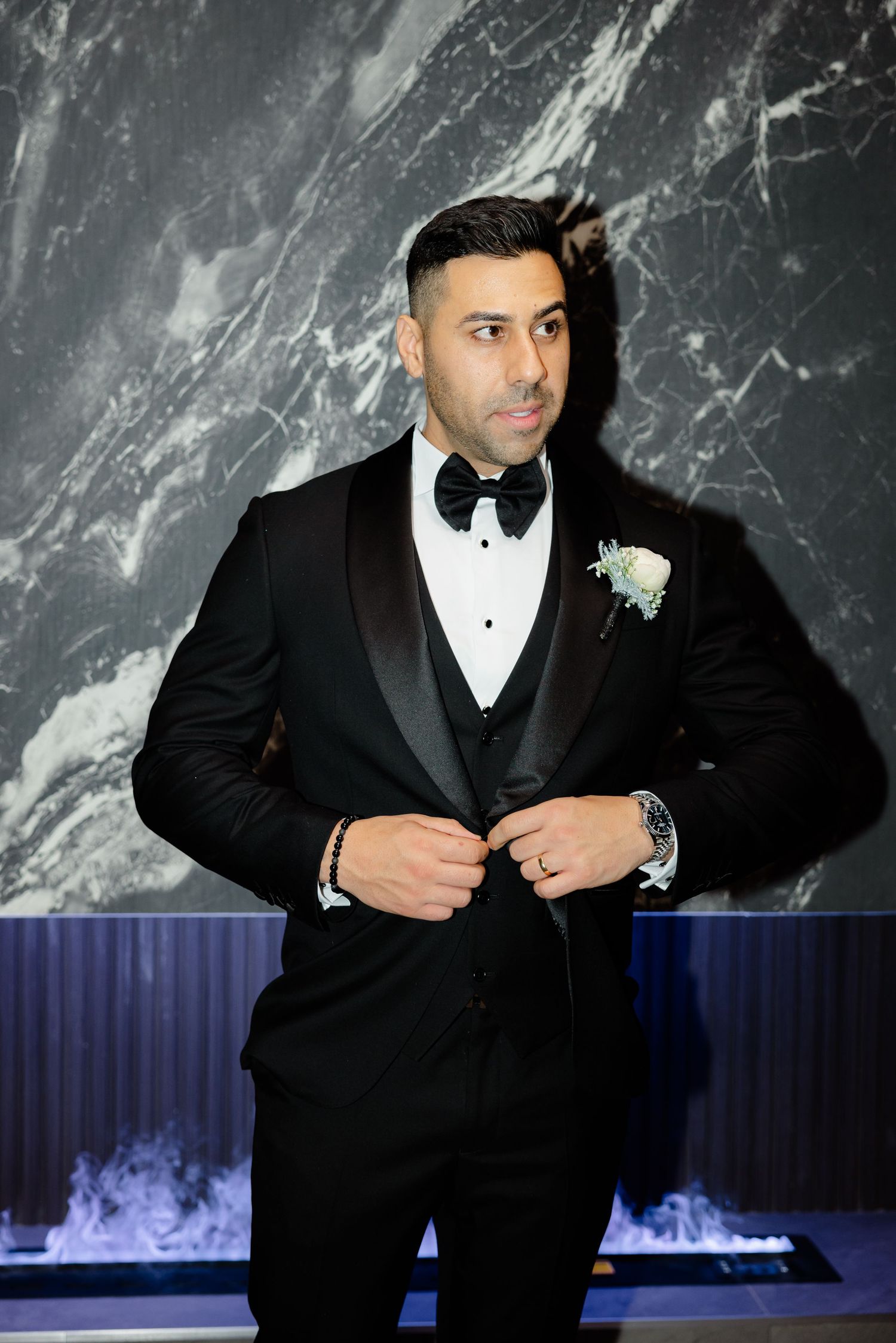 Elegant groom adjusts his tuxedo jacket against dramatic marble and blue-lit background.