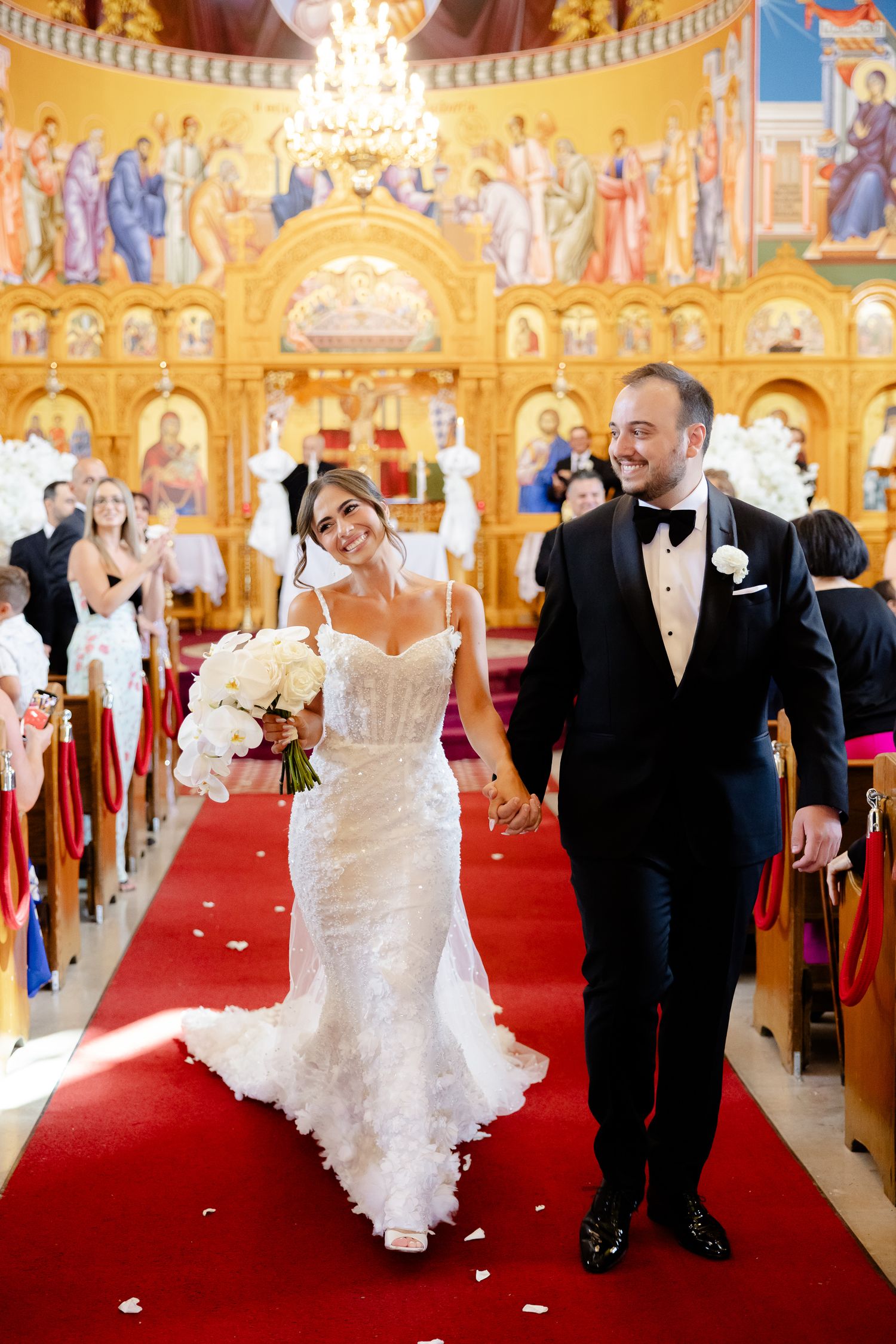 Wedding couple walking and dancing down the red carpet aisle in an ornate Byzantine-style church with golden murals.