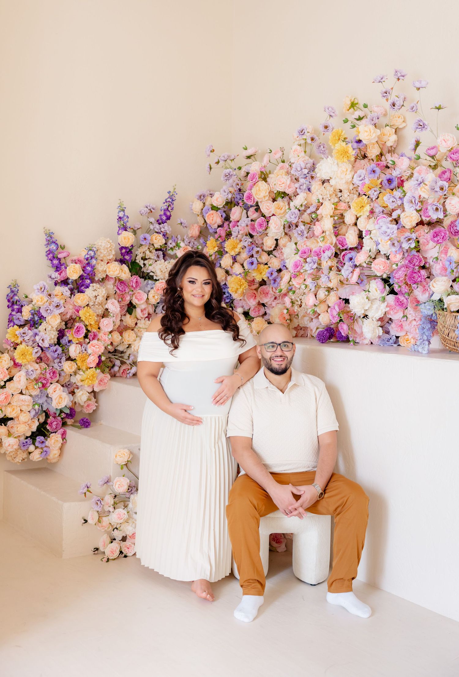 Couple poses against stunning floral backdrop with purple and peach blooms in elegant white and casual attire.