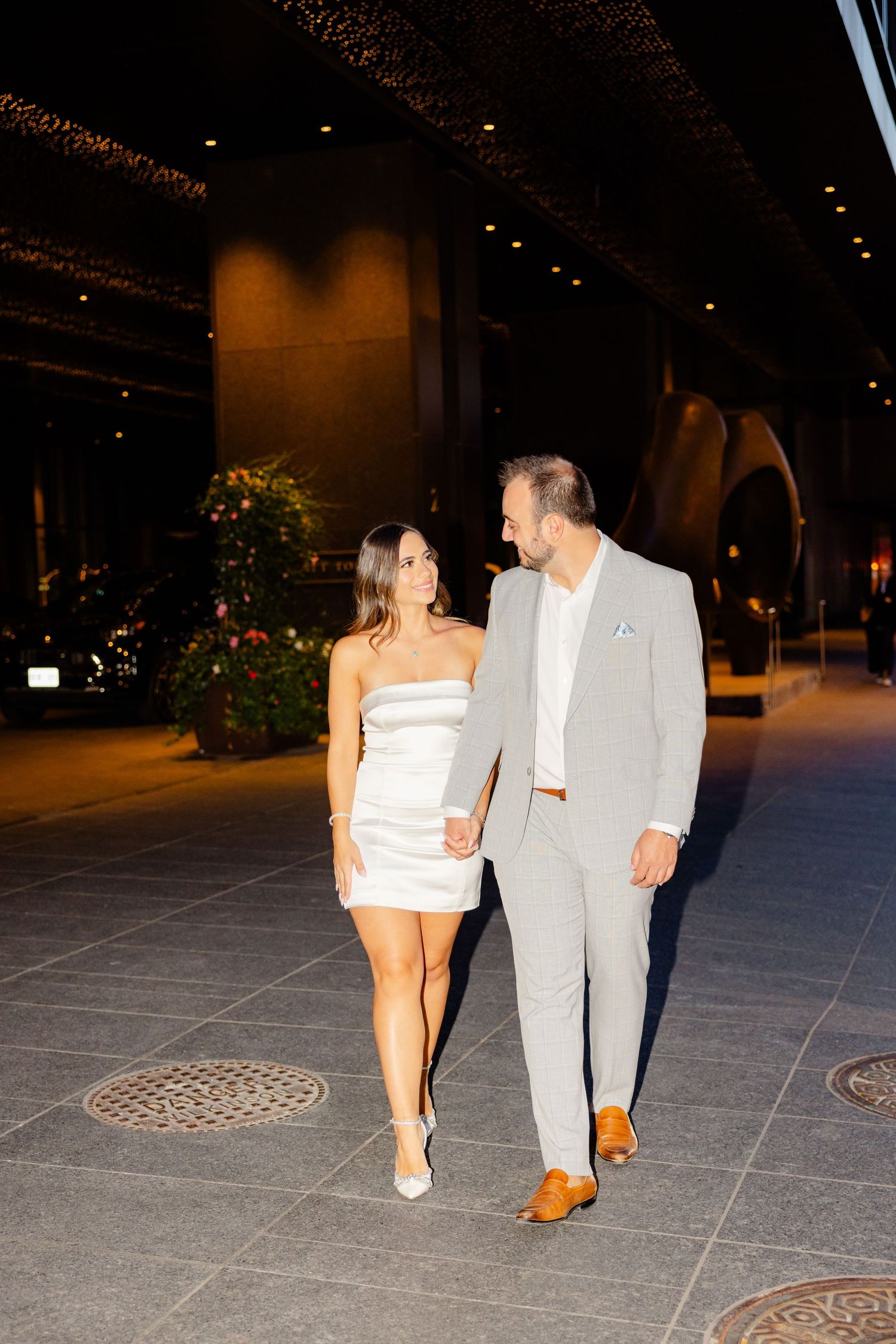 Couple smiles at each other while walking under decorative lighting on city plaza.