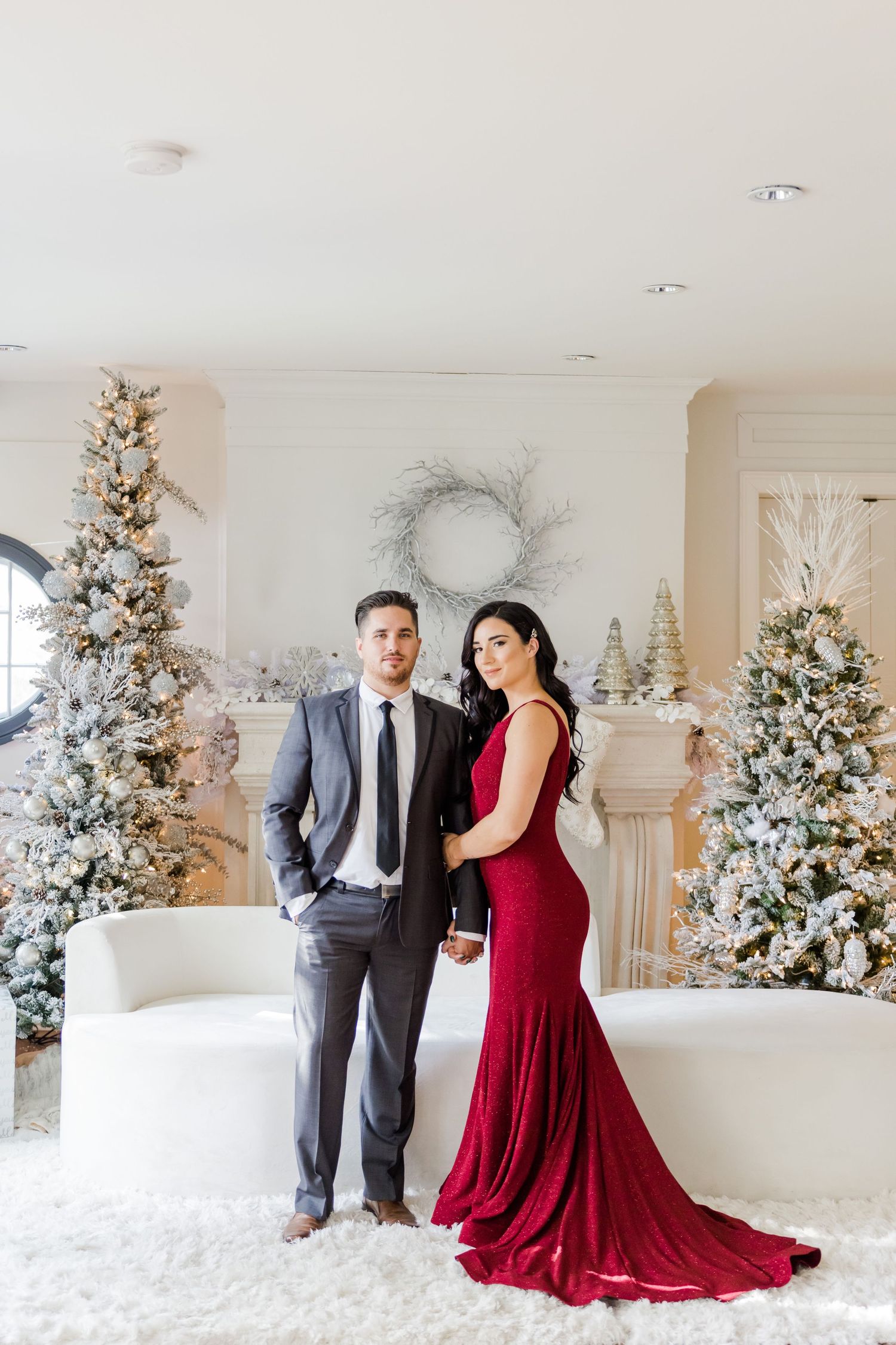 Glamorous formal portrait with red gown and gray suit against a backdrop of snow-flocked Christmas trees.
