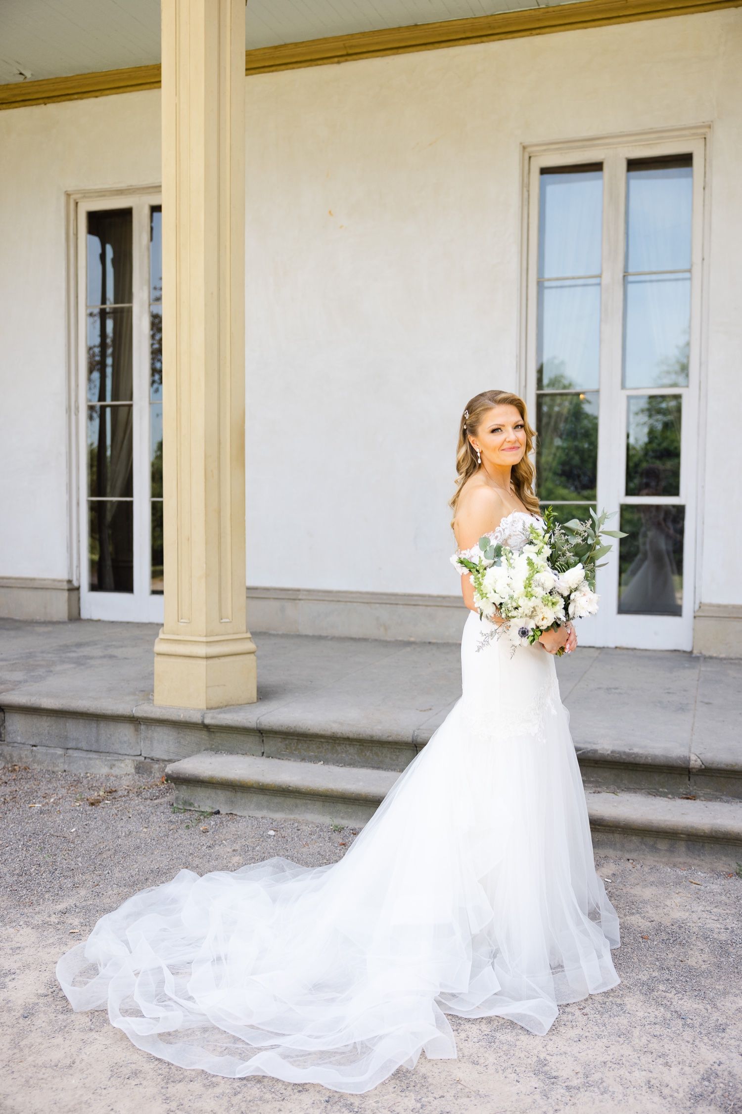 An elegant bride in a fitted white wedding gown with flowing train poses on marble steps by ornate columns.