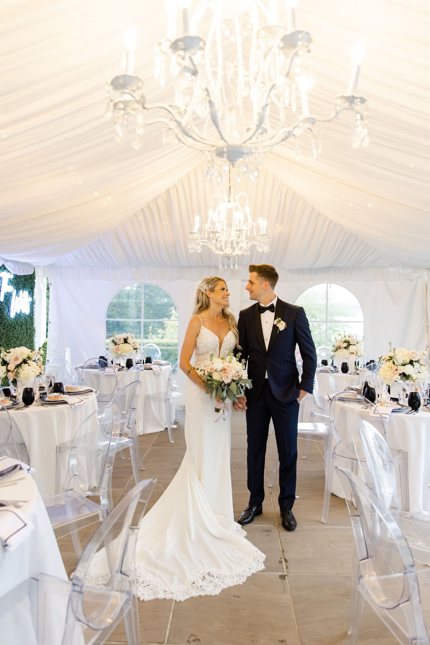 Elegant wedding reception setup in white tent with crystal chandelier and ghost chairs.