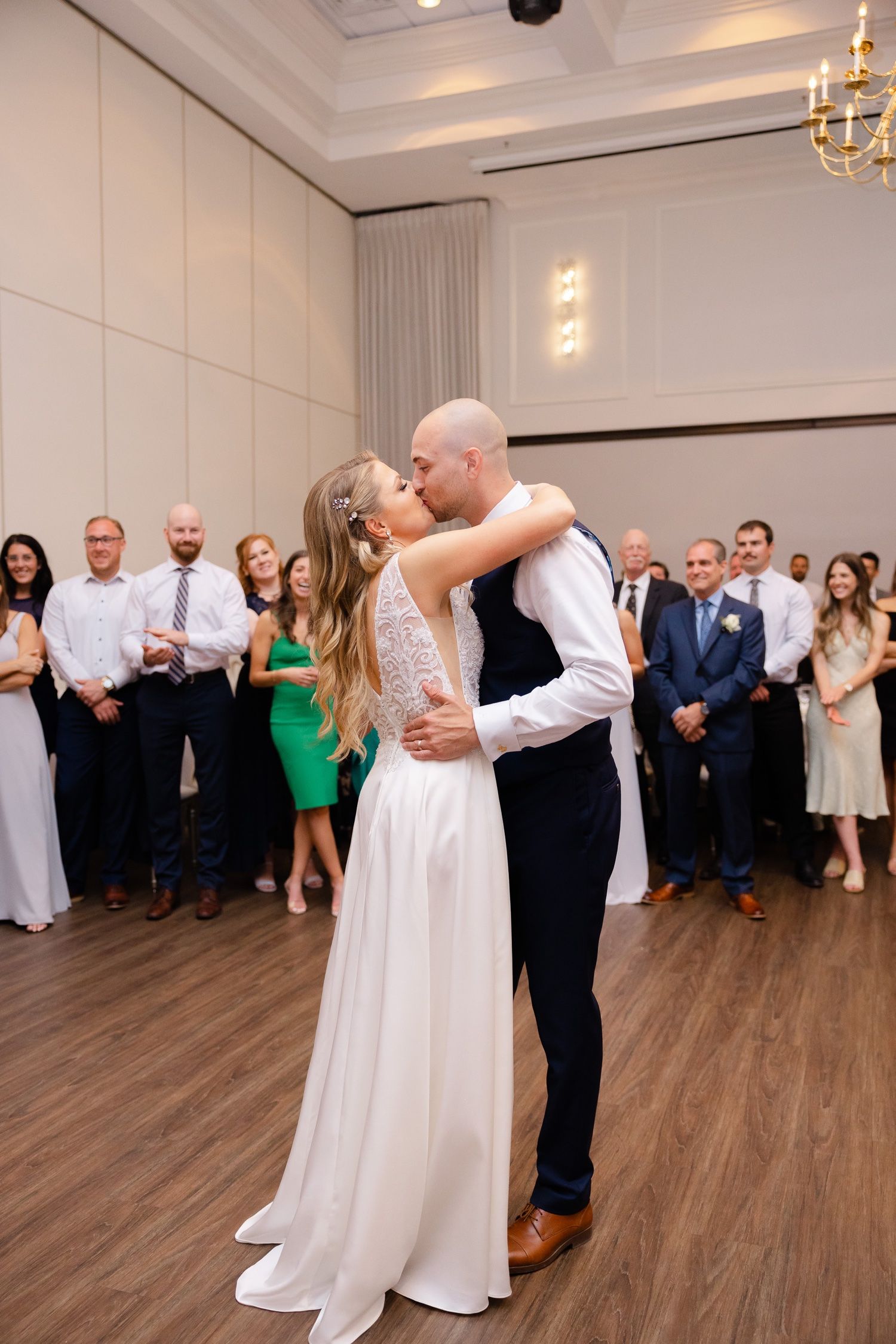Couple embraces during wedding dance as guests watch in elegant ballroom setting.