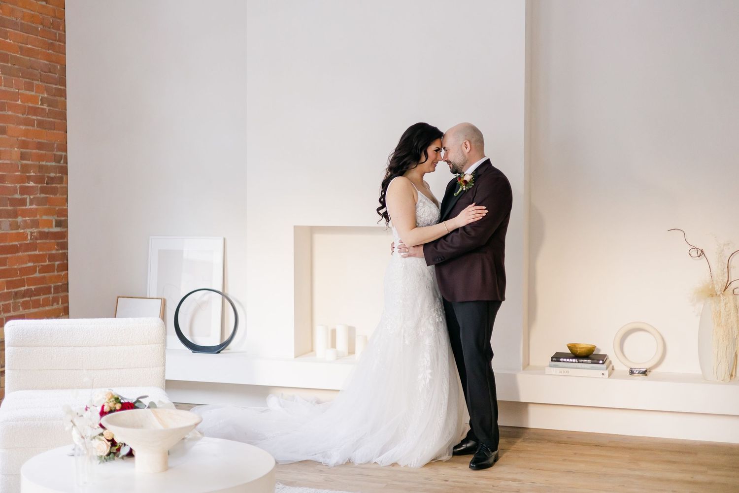 A tender wedding moment captured in a modern white room with brick accent wall and decorative elements.
