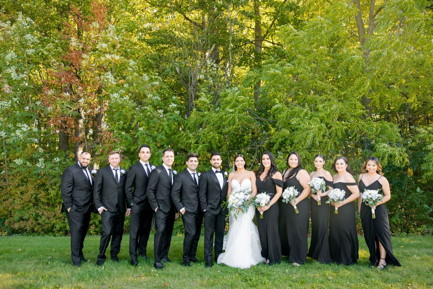 Wedding party in black attire poses together on a grassy lawn with autumn trees in the background.