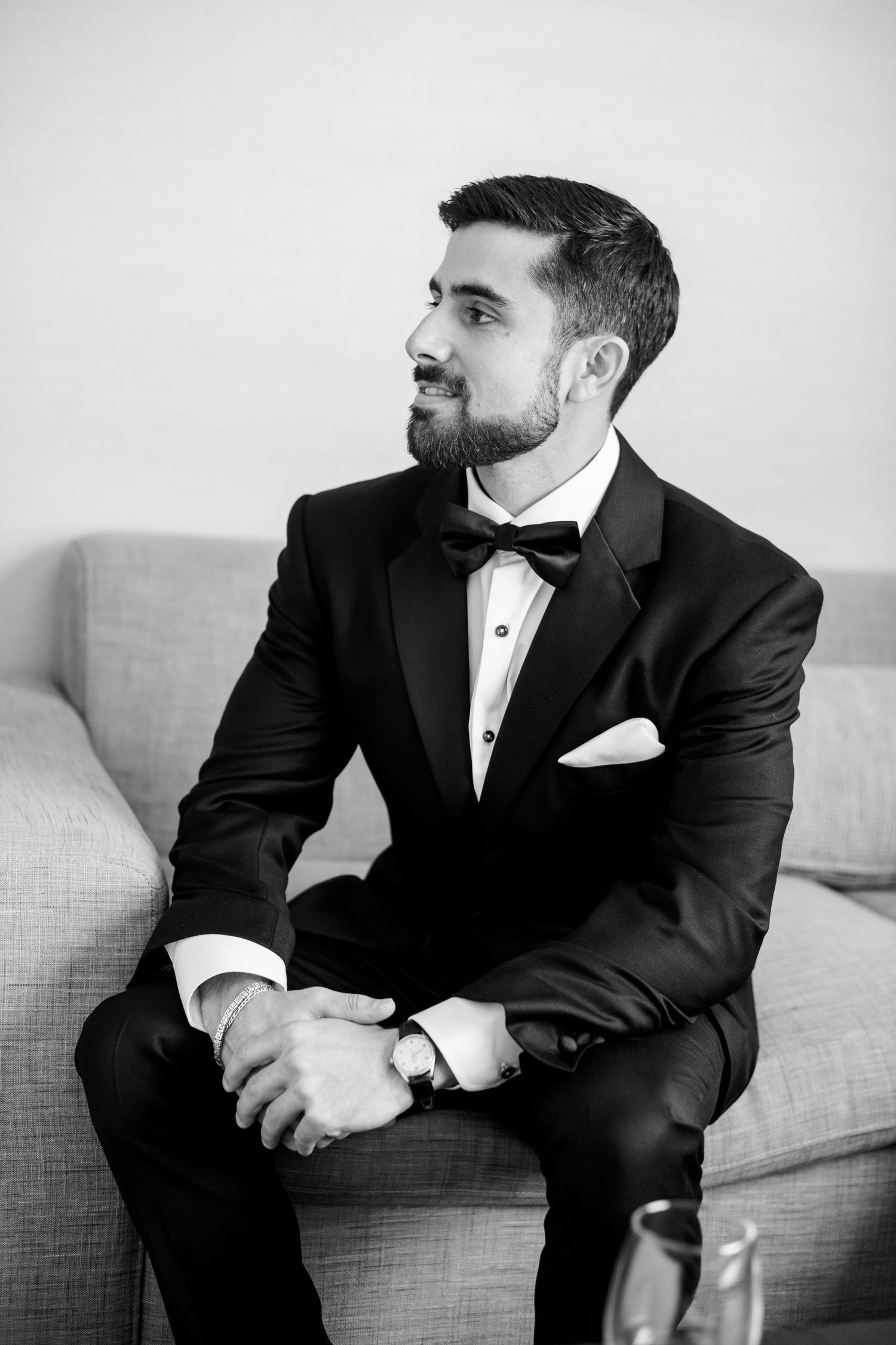 A groom in a classic black tuxedo and bow tie sits contemplatively on a grey couch.