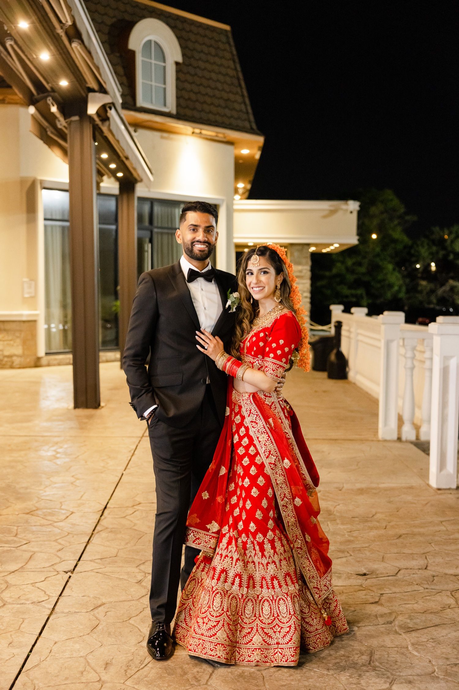 Couple in formal wedding attire poses outside illuminated venue at night.