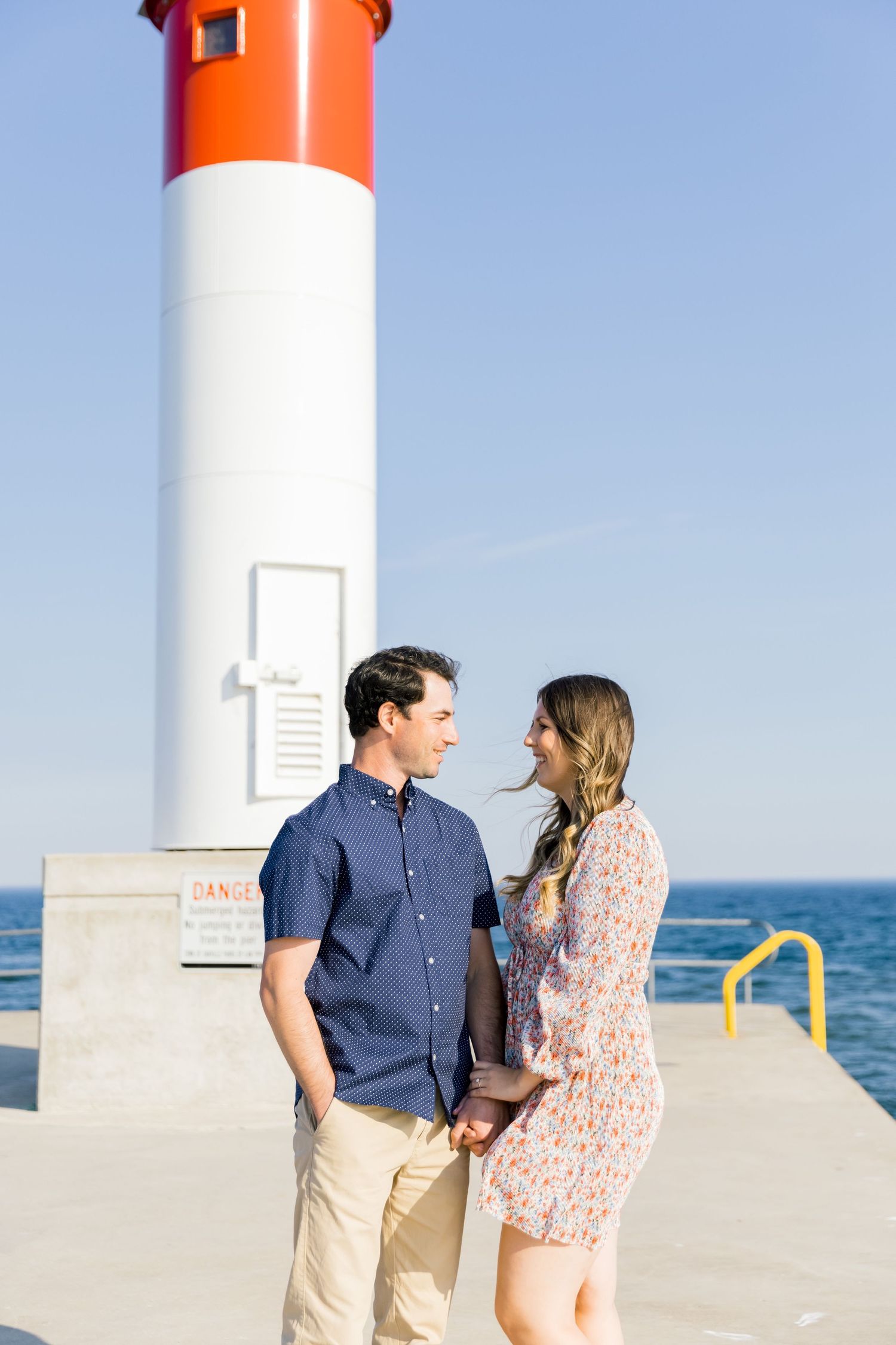 A couple shares an intimate moment beside a tall lighthouse with orange top on a sunny day.