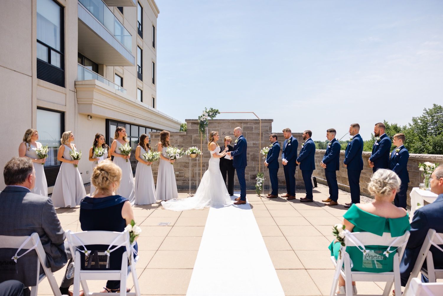 Wide shot of a rooftop wedding ceremony with guests seated and the wedding party in navy and blush colors.