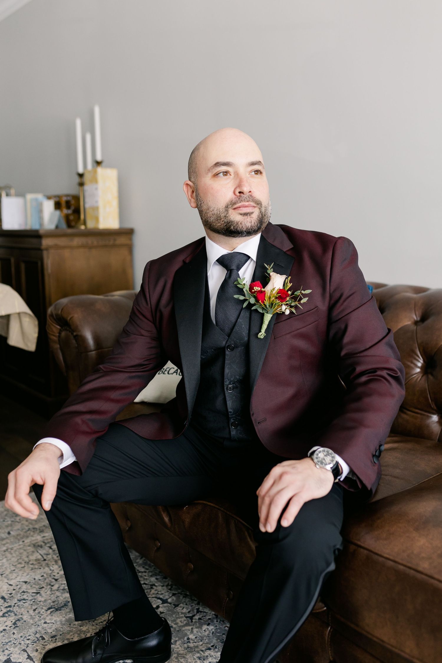Man in burgundy suit jacket and black tie sitting on brown leather couch with red boutonniere.