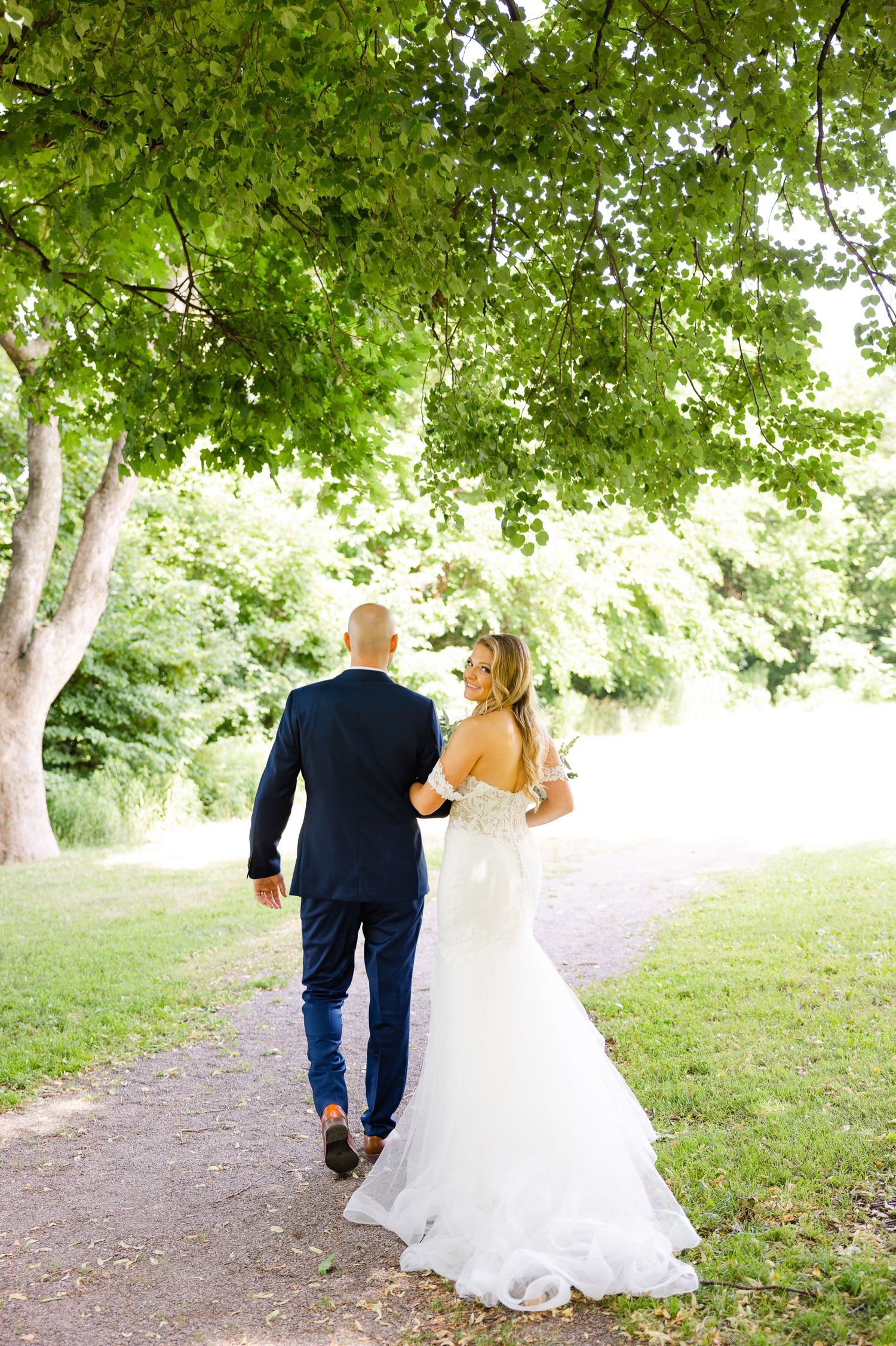 Couple walking down tree-lined path during summer wedding celebration.