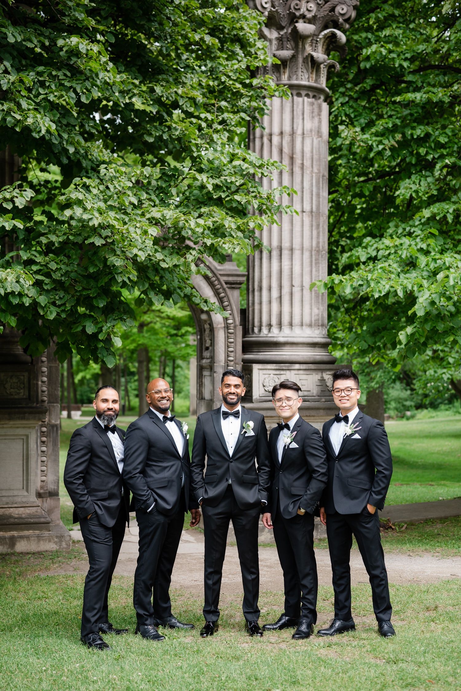 Groomsmen in classic black tuxedos pose by ornate stone column in garden setting.