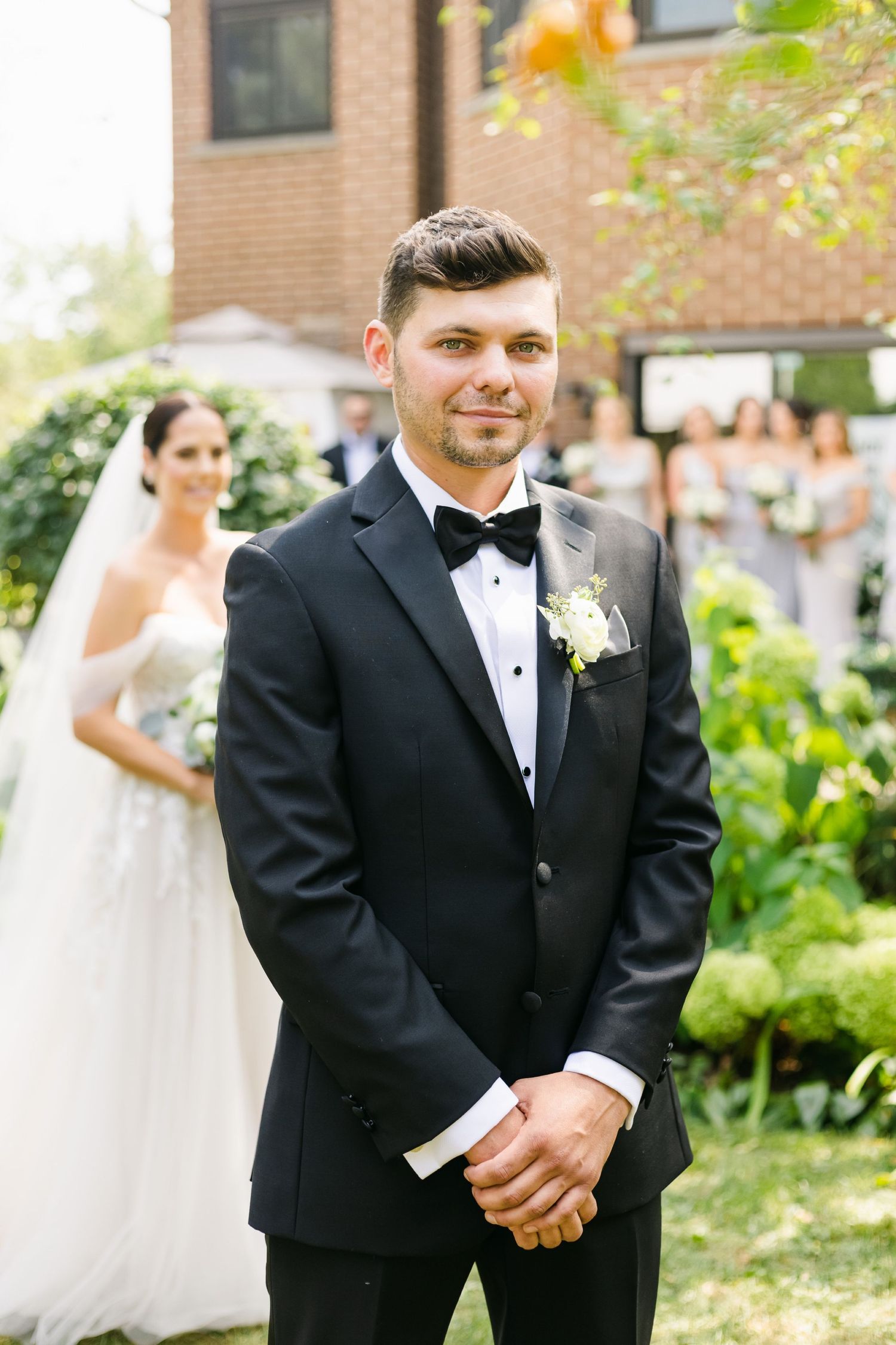 Formal wedding attire with black tuxedo and white boutonniere in garden setting.