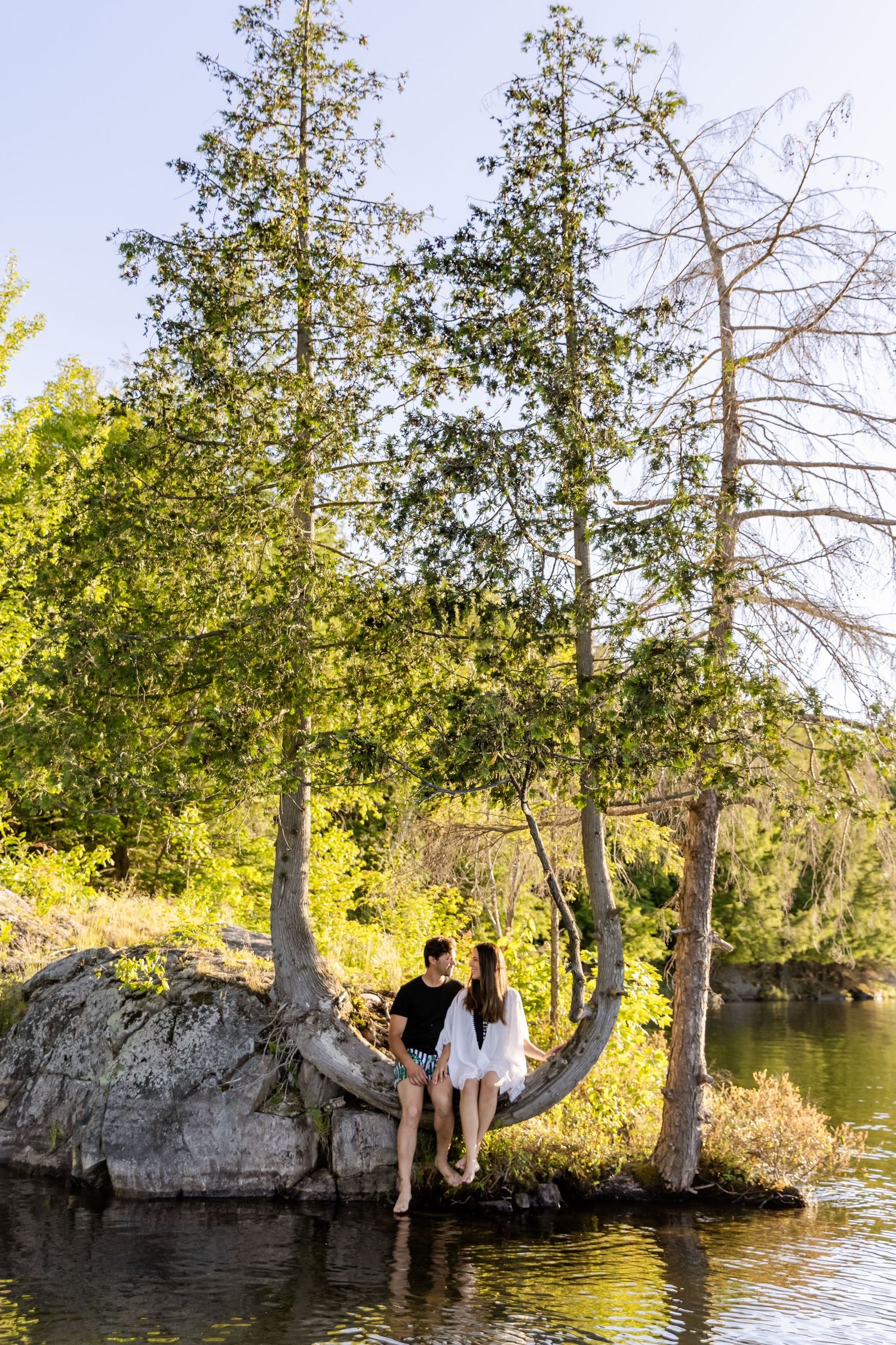 Couple sitting together on a rock formation by a serene lake surrounded by tall trees in golden sunlight.