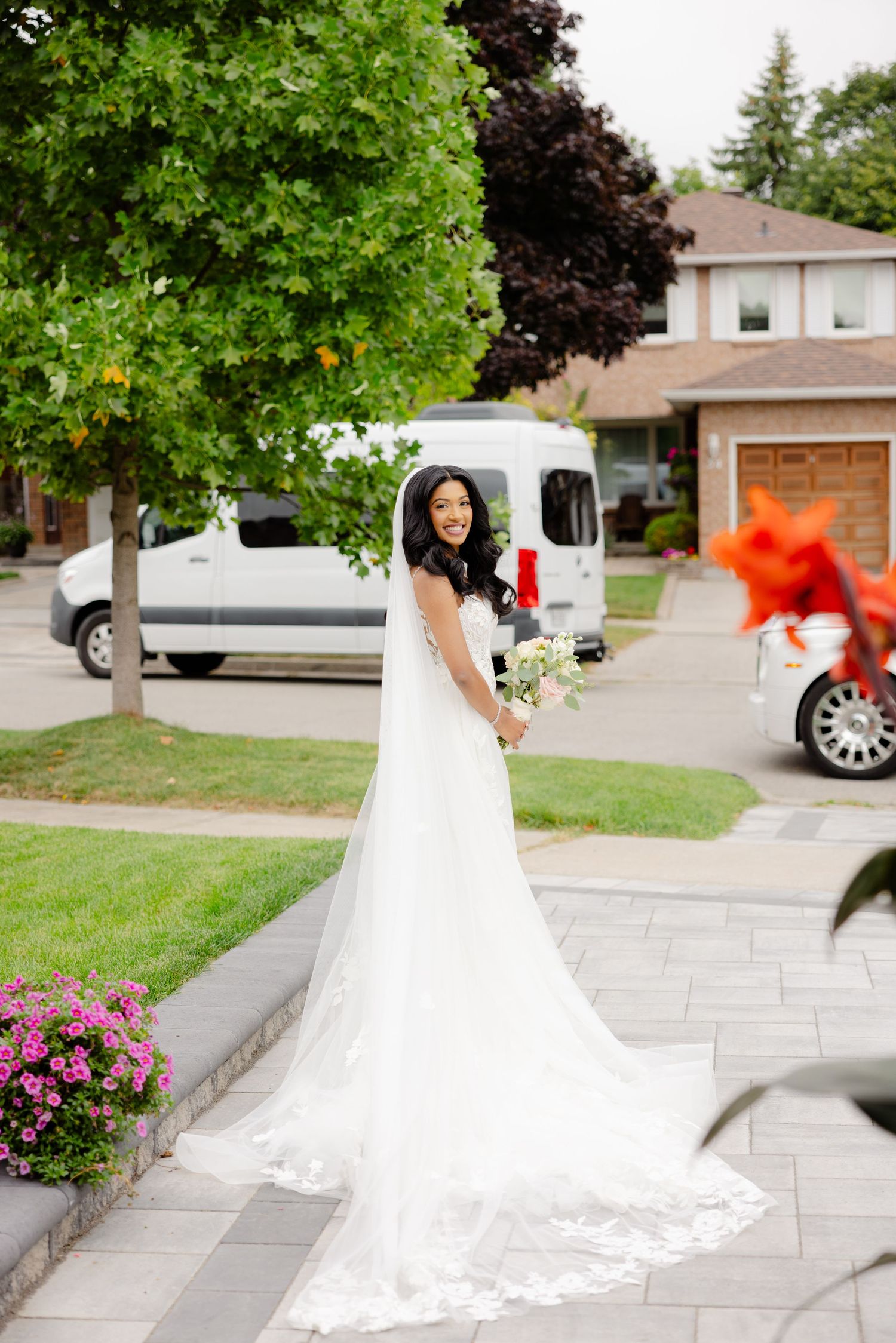Bride in flowing white gown looks over shoulder on walkway with landscaped garden.