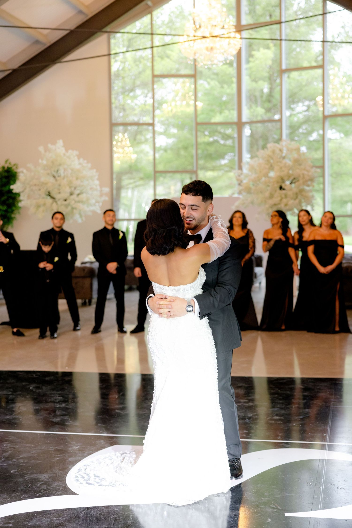 Couple shares first dance in elegant ballroom with bridal party watching from background.