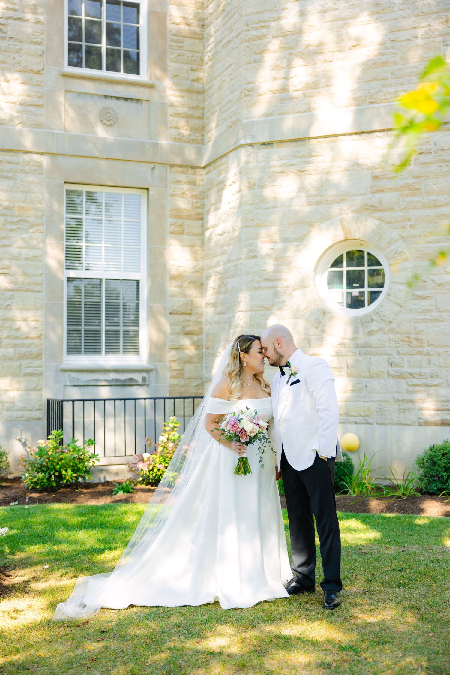 A wedding couple shares an intimate moment in front of a stone building with round window.
