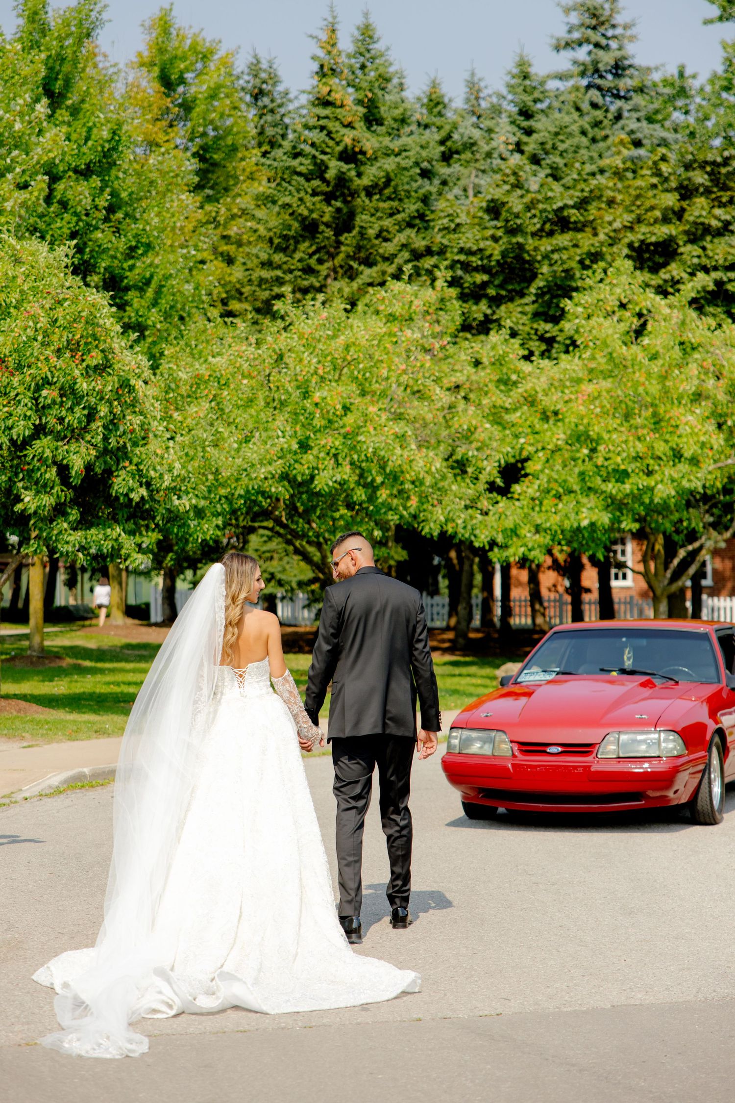 Wedding couple walks away from their red car with beautiful green trees in background.