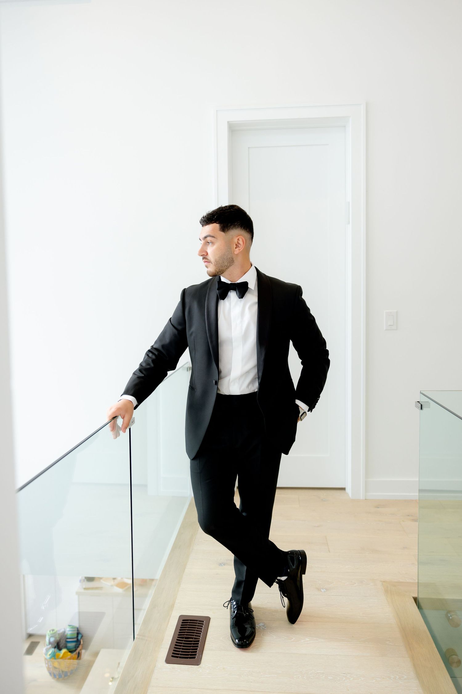 Stylish groom leans against glass railing in modern white hallway wearing formal black tuxedo.