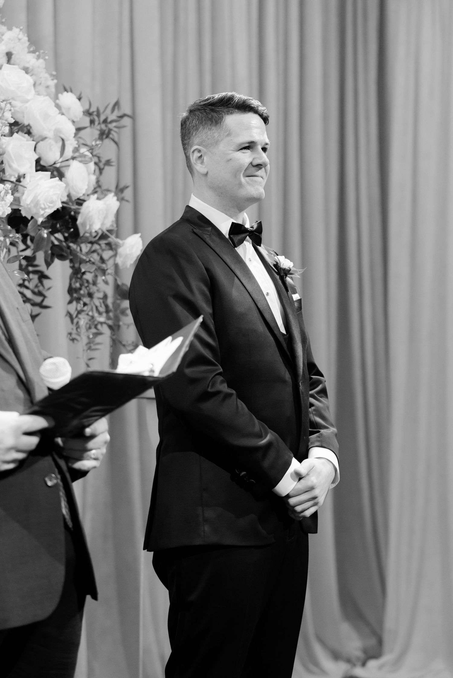 Groom in classic black tuxedo and bowtie smiles during wedding ceremony.