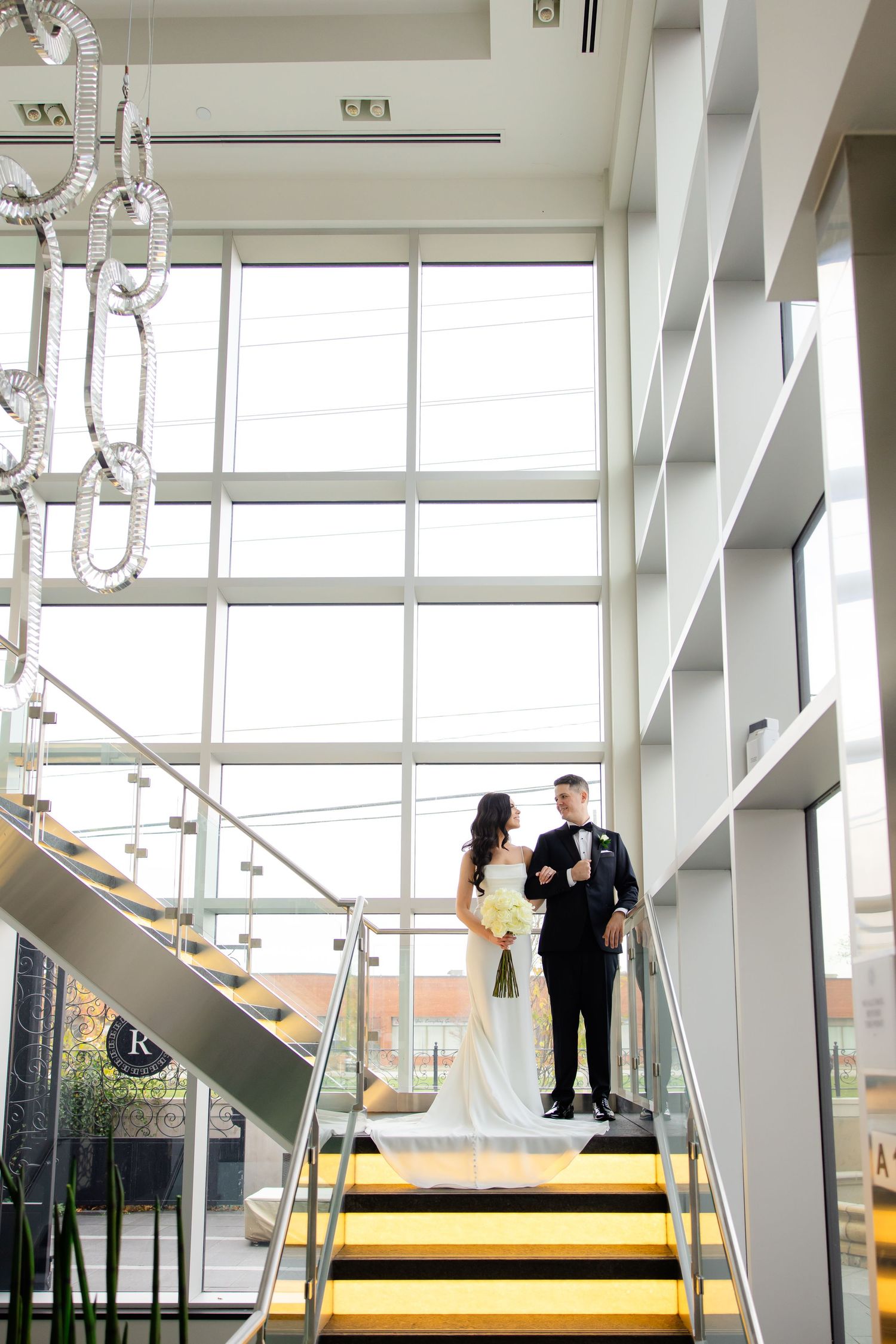 Wedding photo captured on modern staircase with chandelier and floor-to-ceiling windows.