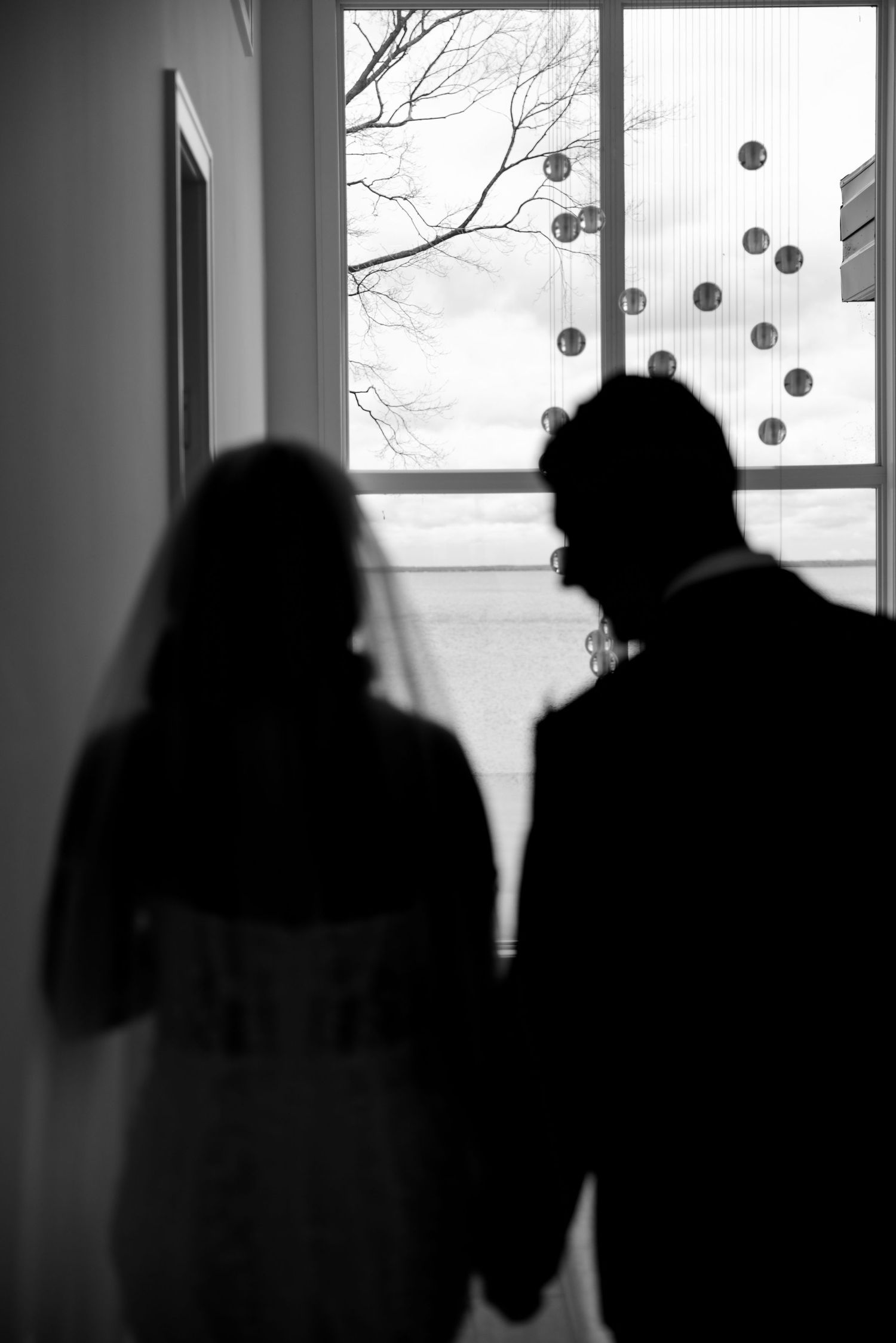 Silhouetted couple admires hanging decorations through window in romantic black and white photo.