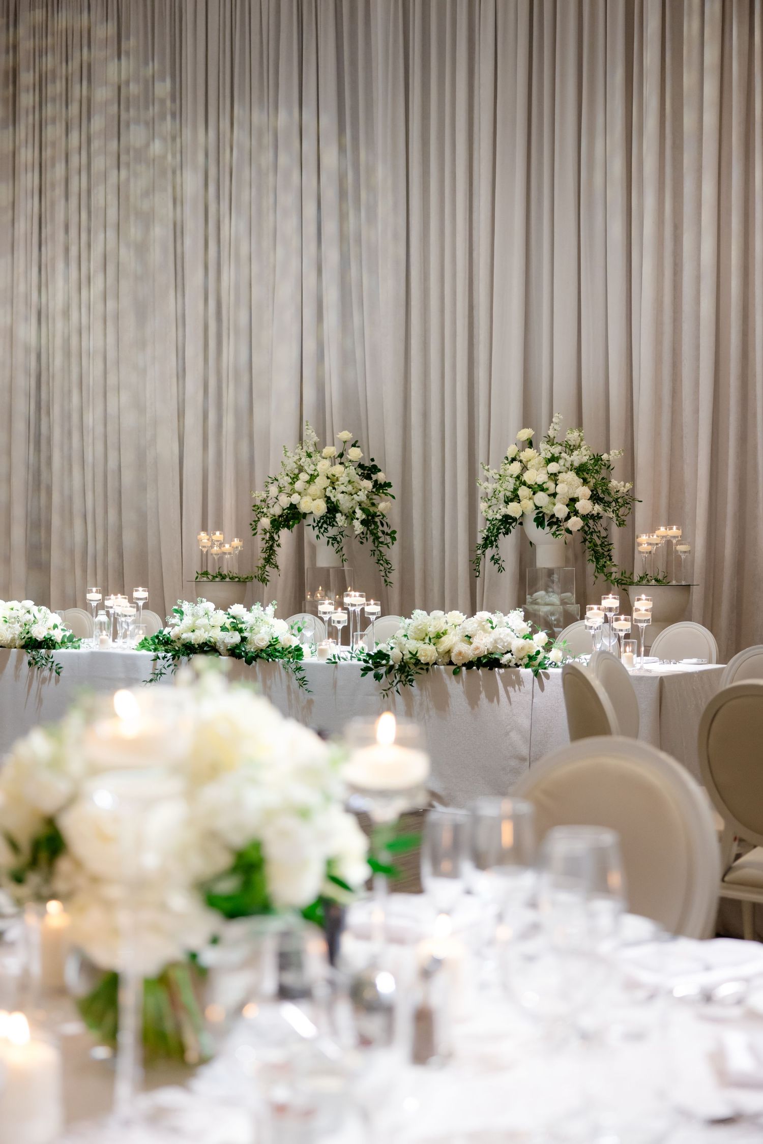 Wedding reception room with draped curtains and white floral arrangements on round tables.