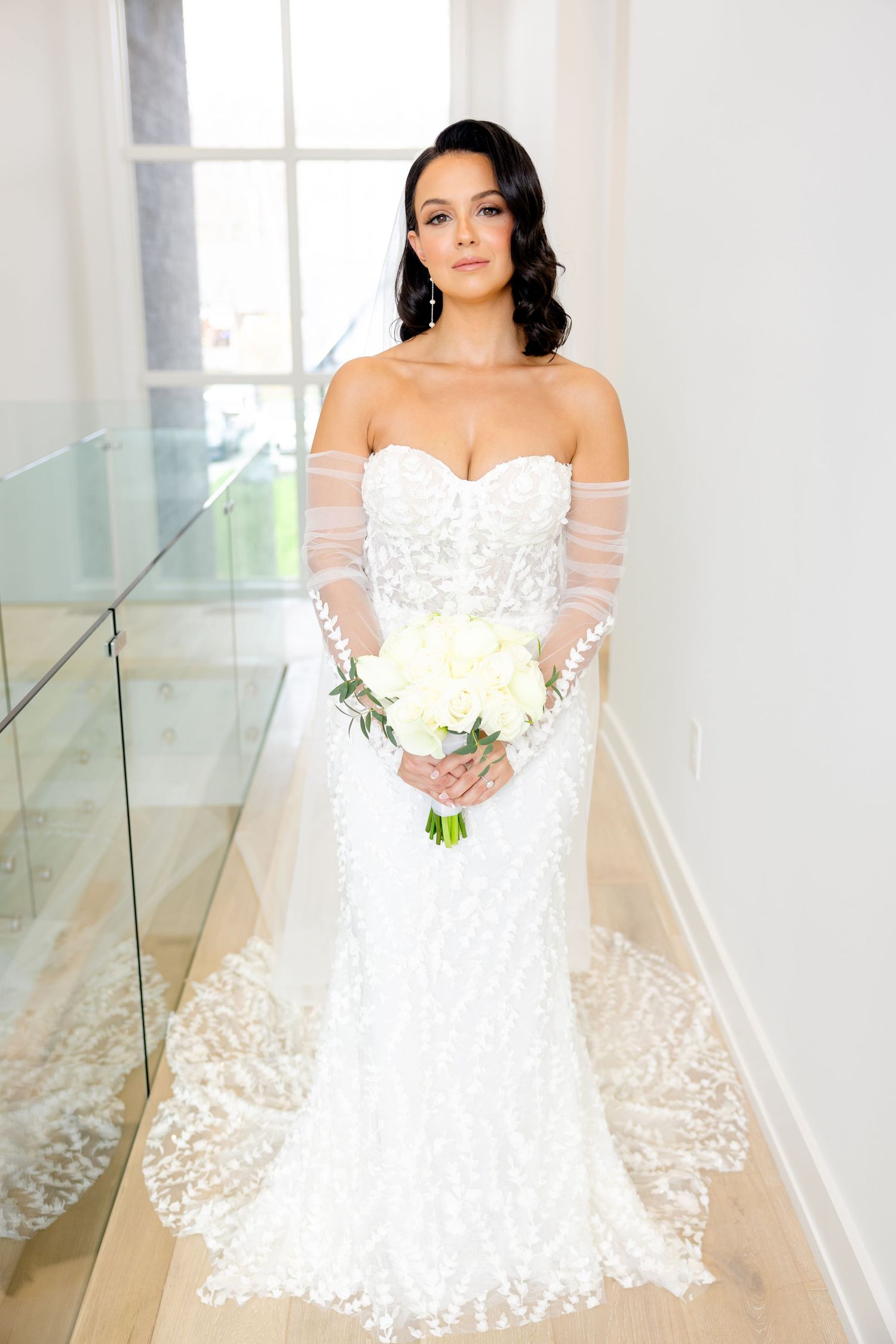 Full length portrait of bride in off-shoulder lace gown holding cream roses in modern hallway.