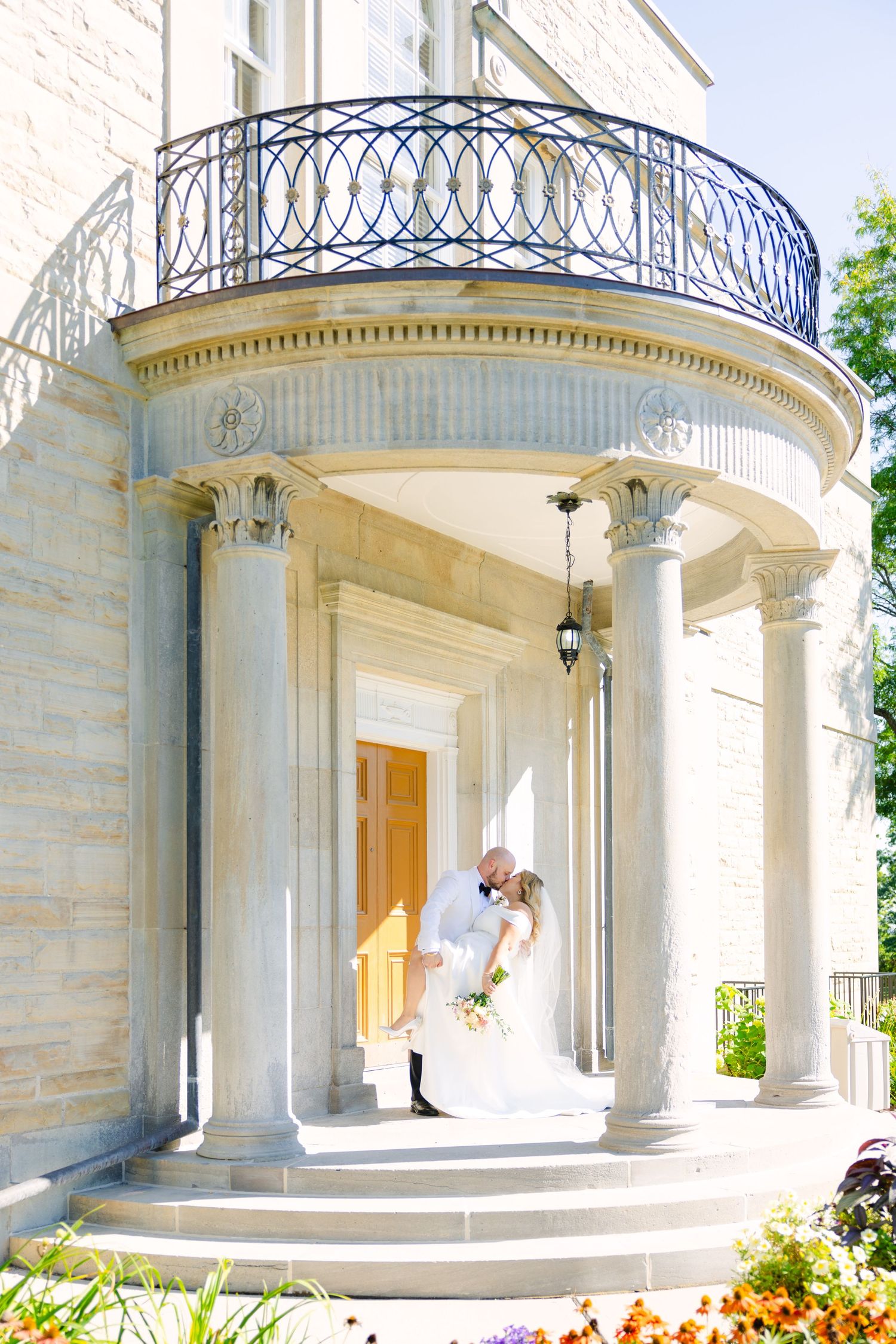 Elegant stone portico with ornate iron railing features a bright orange door and classic architectural details.