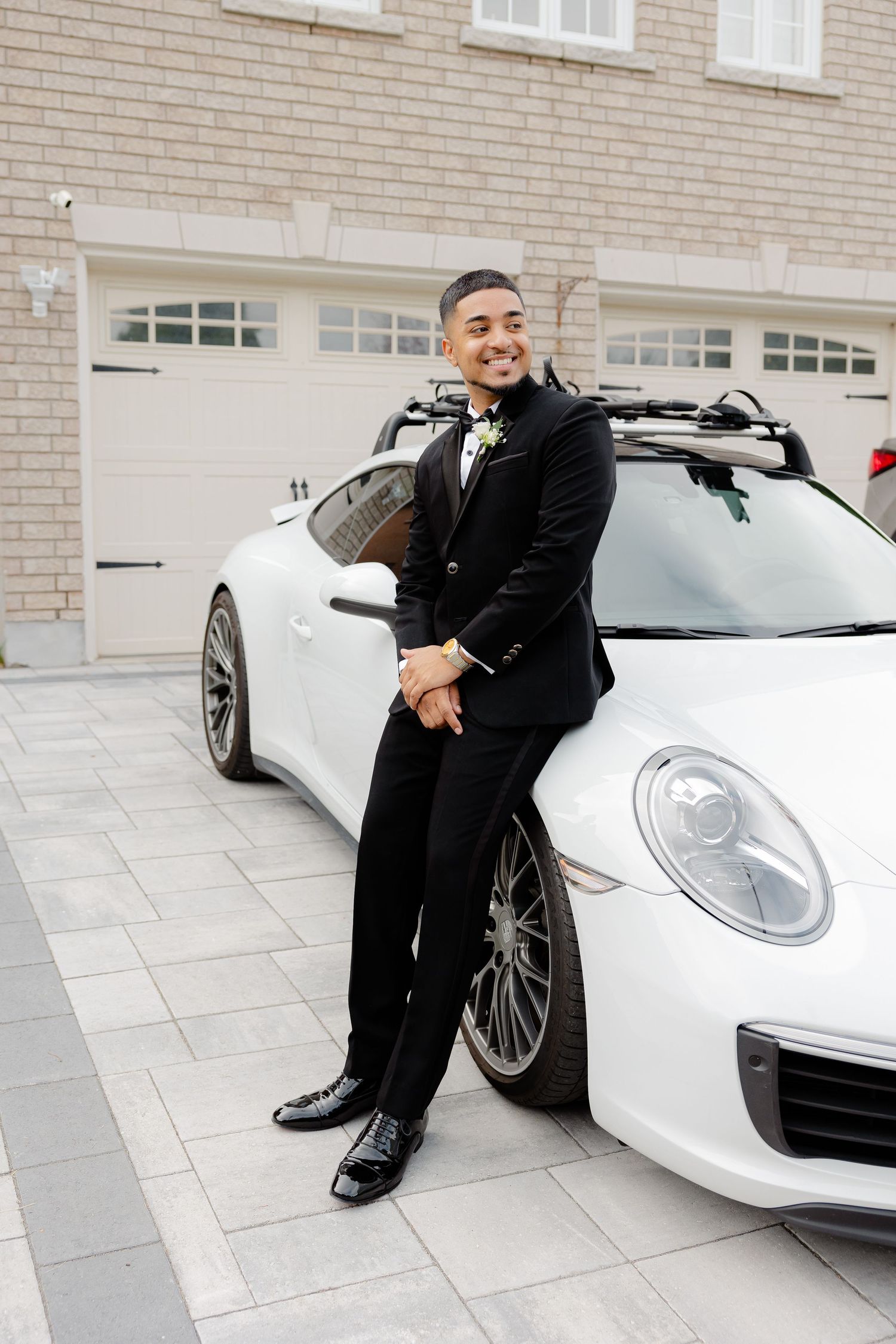 Formal portrait of someone in a tuxedo leaning against a white Porsche sports car in front of a garage.