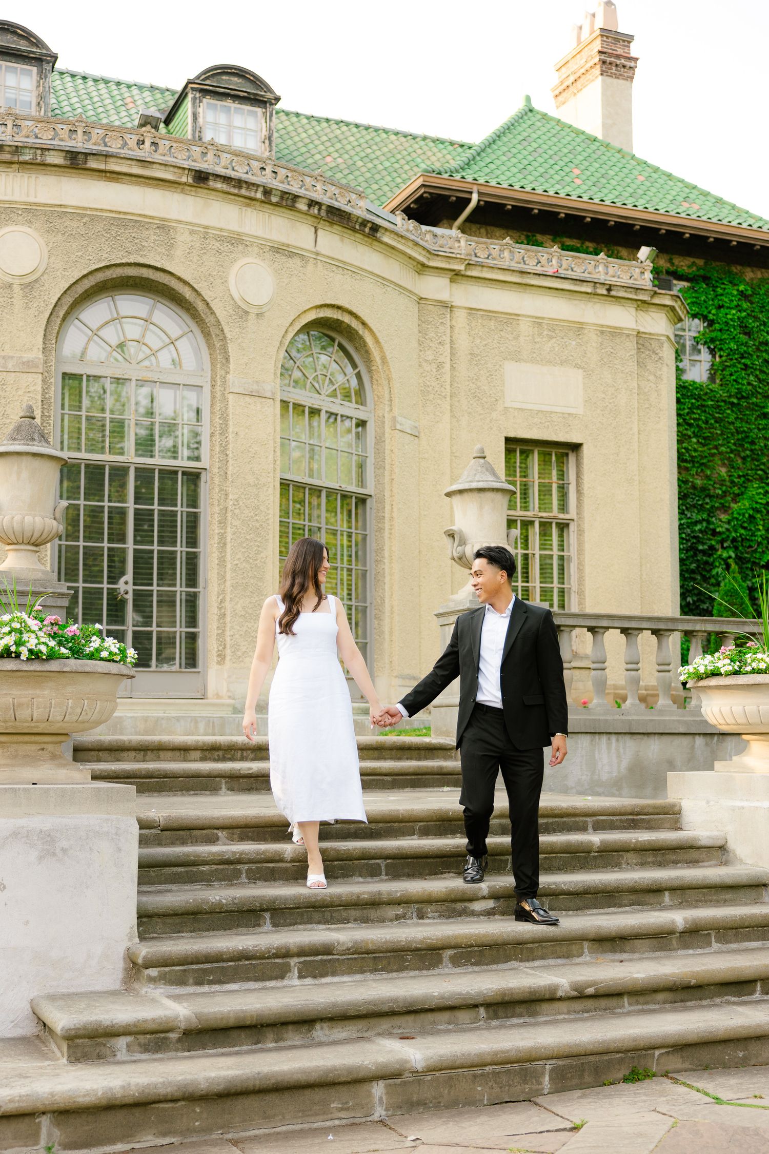 A couple descending grand stone steps at a historic mansion with arched windows and ivy-covered walls.