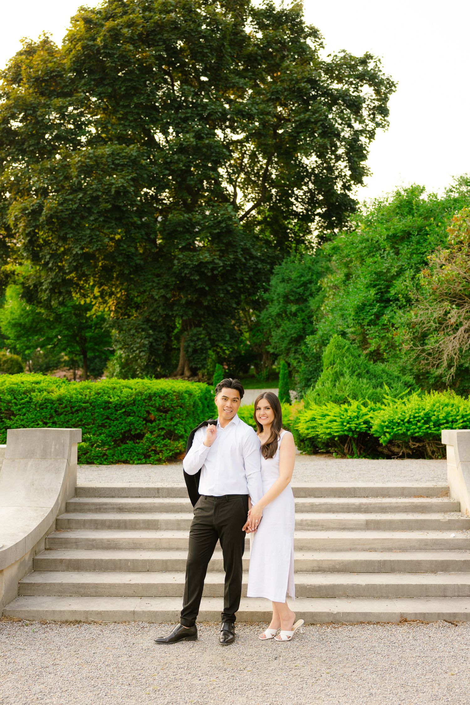 Couple in formal wear poses on stone steps in a lush garden setting with mature trees in the background.