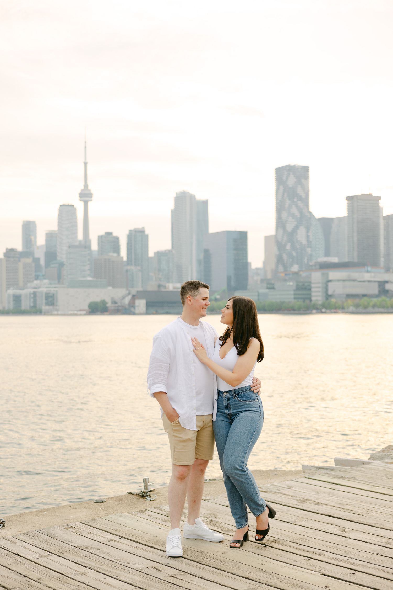 Couple embracing on wooden dock with Toronto's downtown skyline visible across the water.