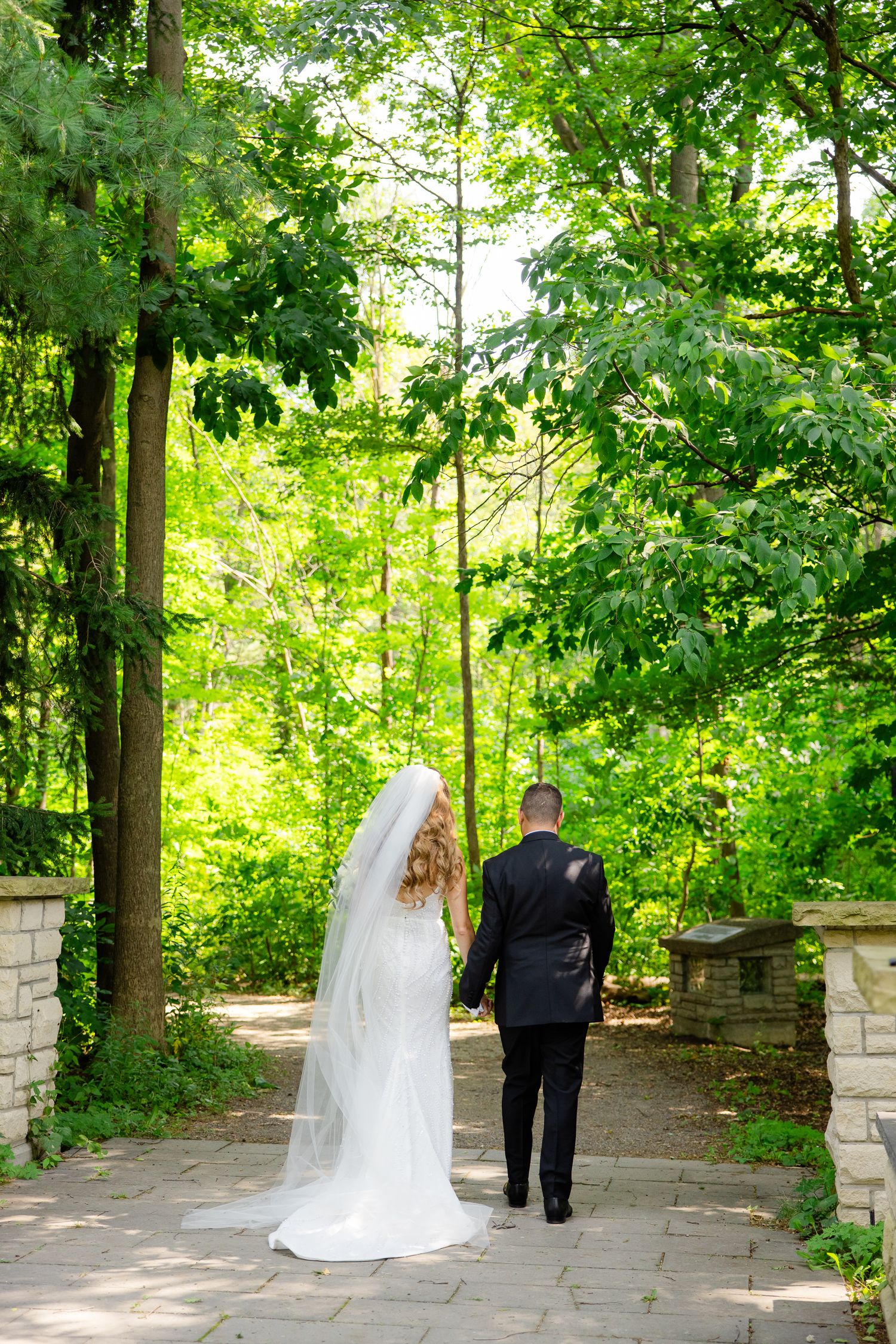 Wedding couple walking away down a garden path with stone walls and green forest backdrop.