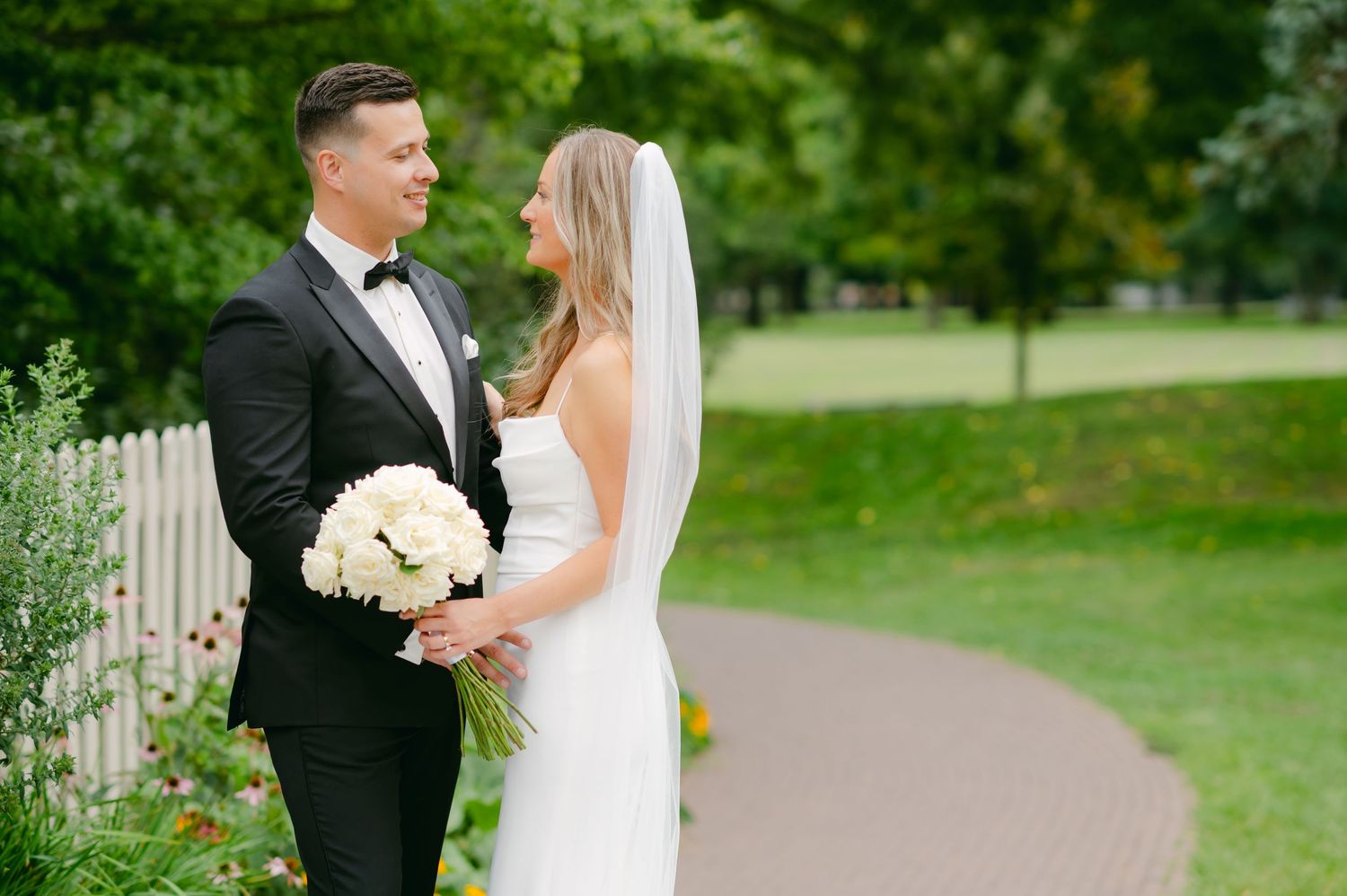 Newlywed couple stands together on garden path with white picket fence and green foliage in background.