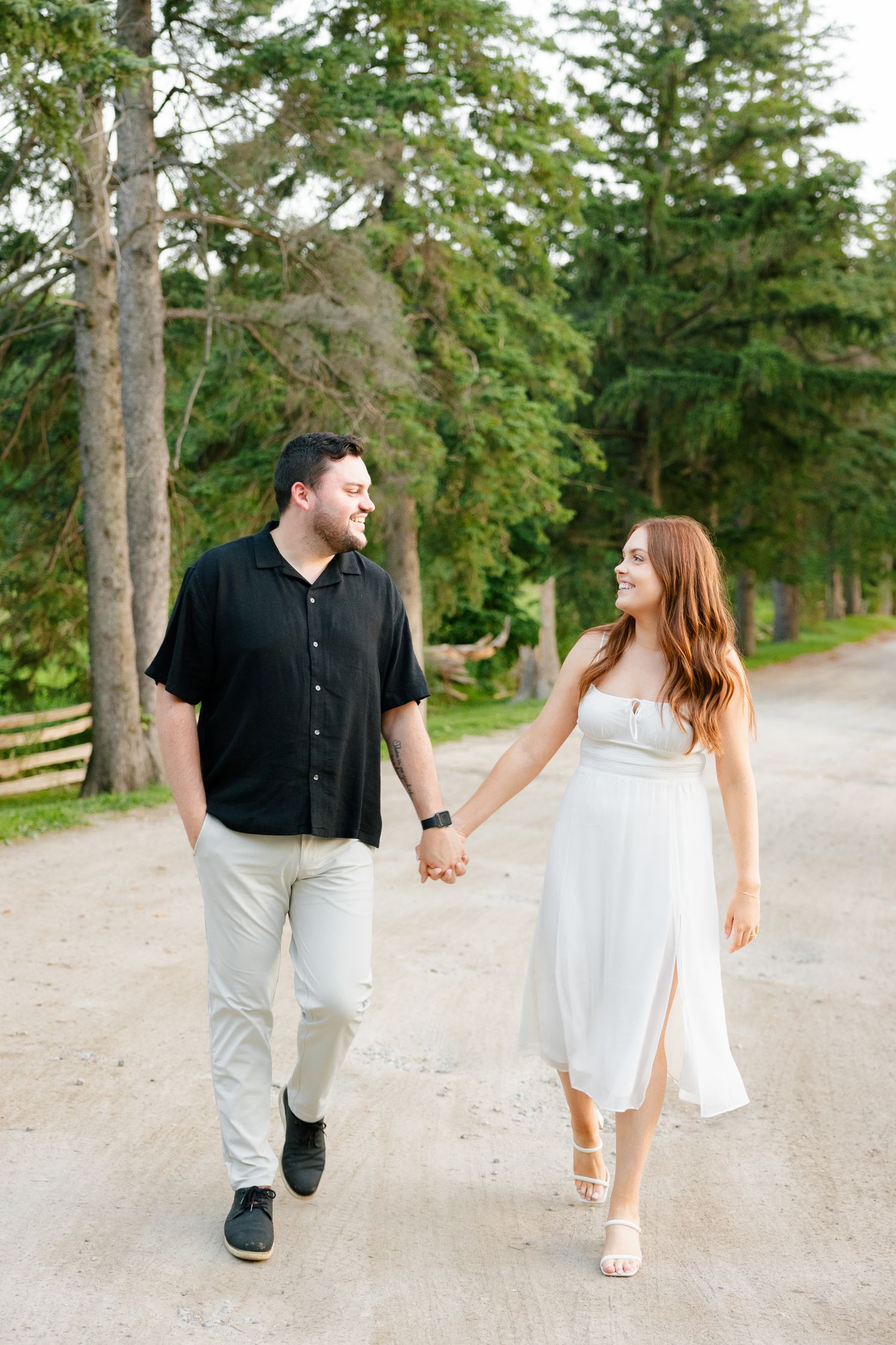A couple strolls together on a peaceful dirt path lined with evergreen trees.