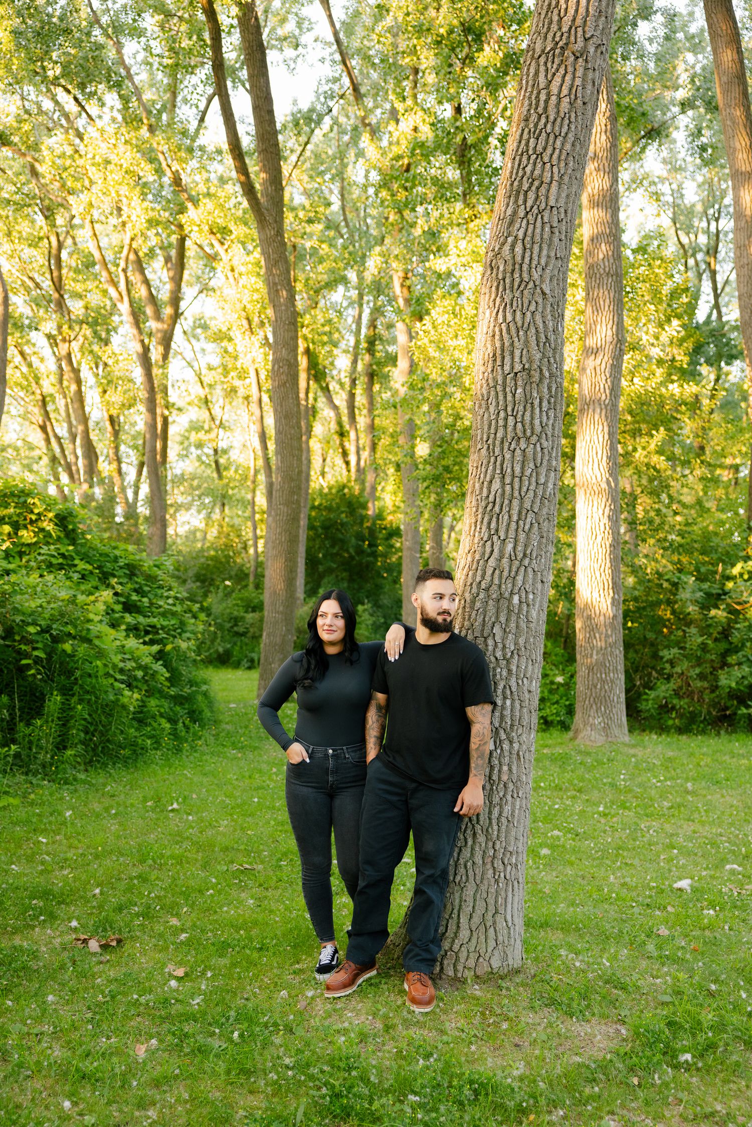 Couple in matching black outfits standing by tall trees in sunny woodland setting.