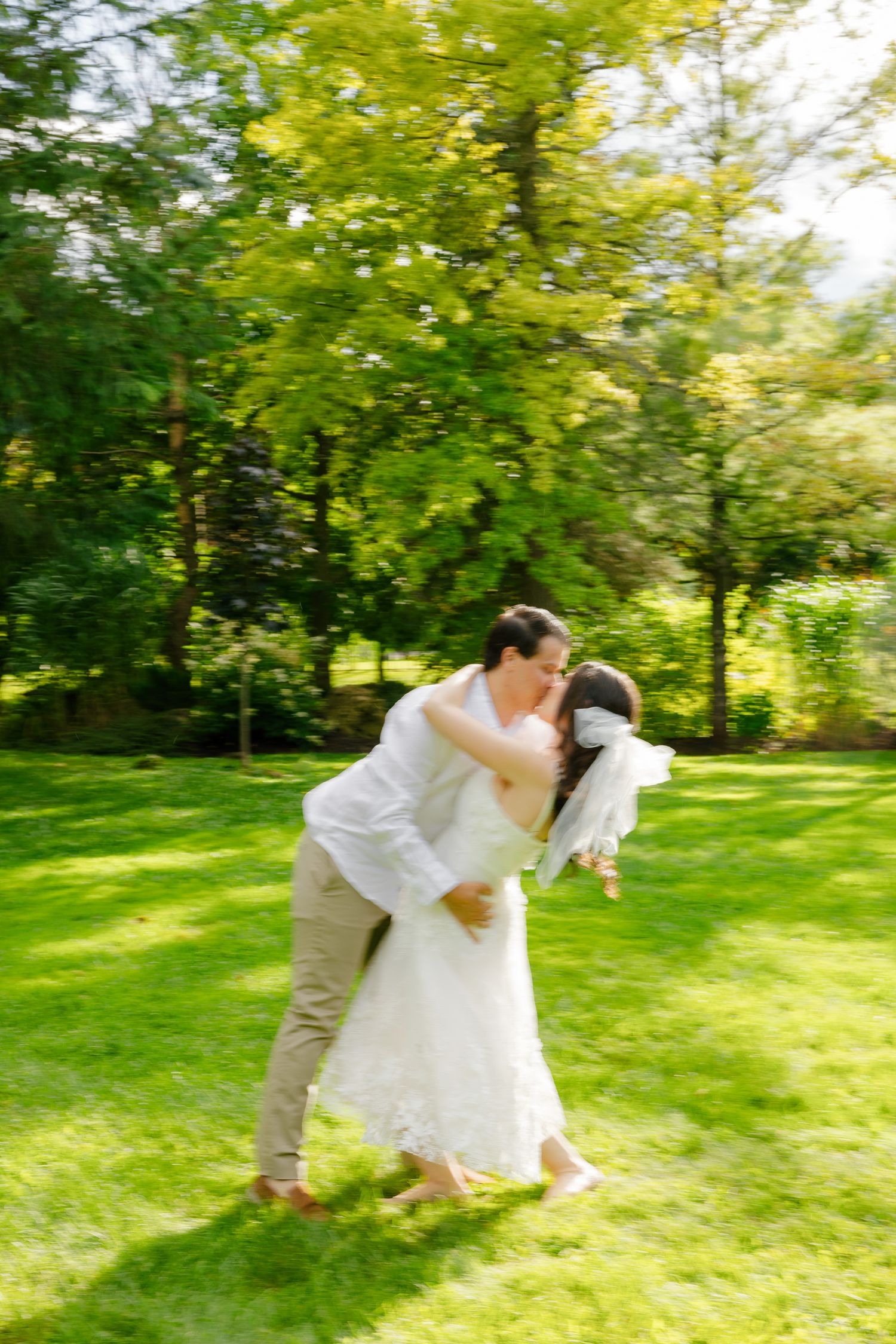 A couple in wedding attire performs a romantic dip pose in a sunlit garden.
