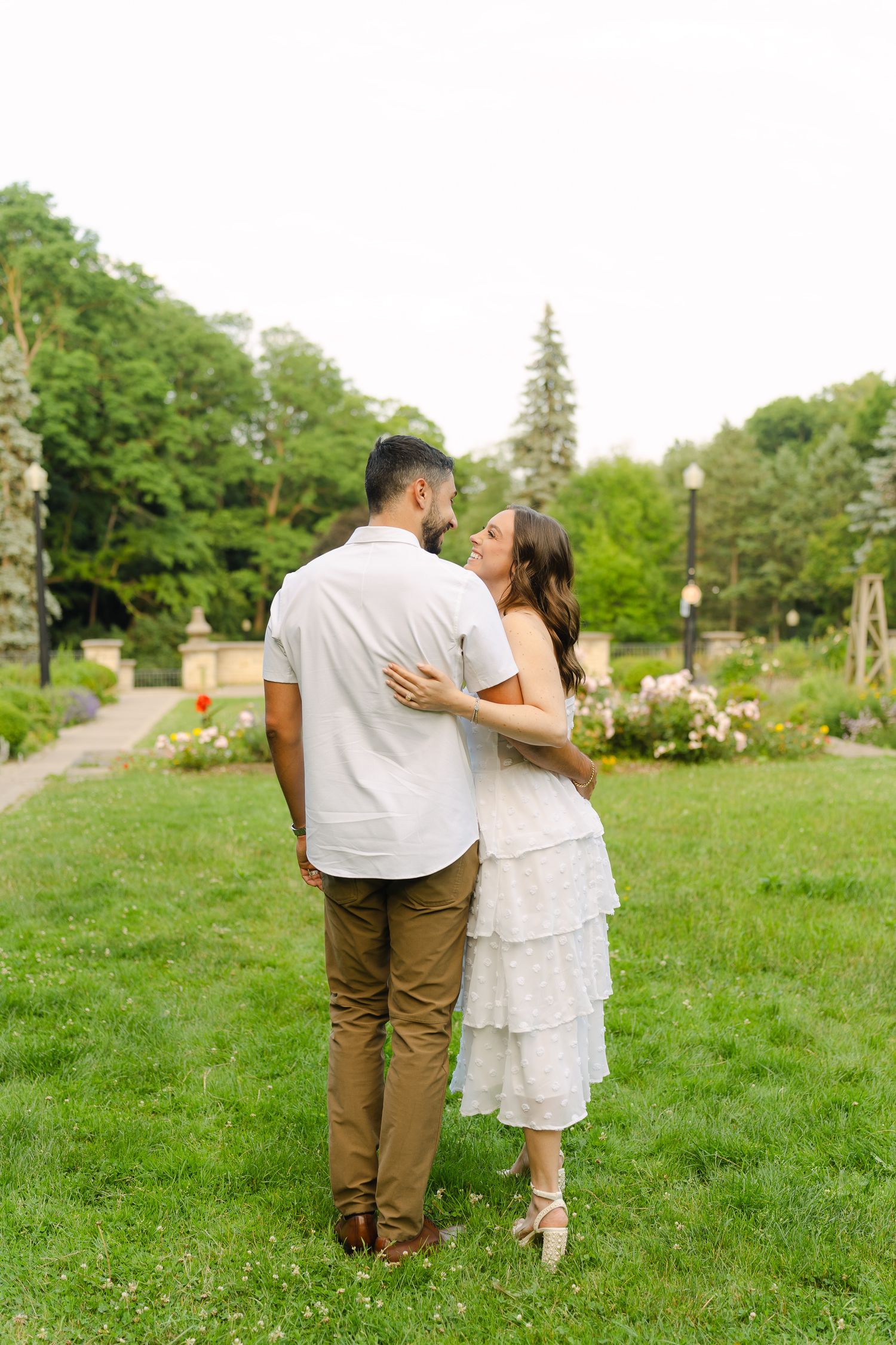 A couple embraces while standing on a lawn with flowering gardens and pathways in the background.