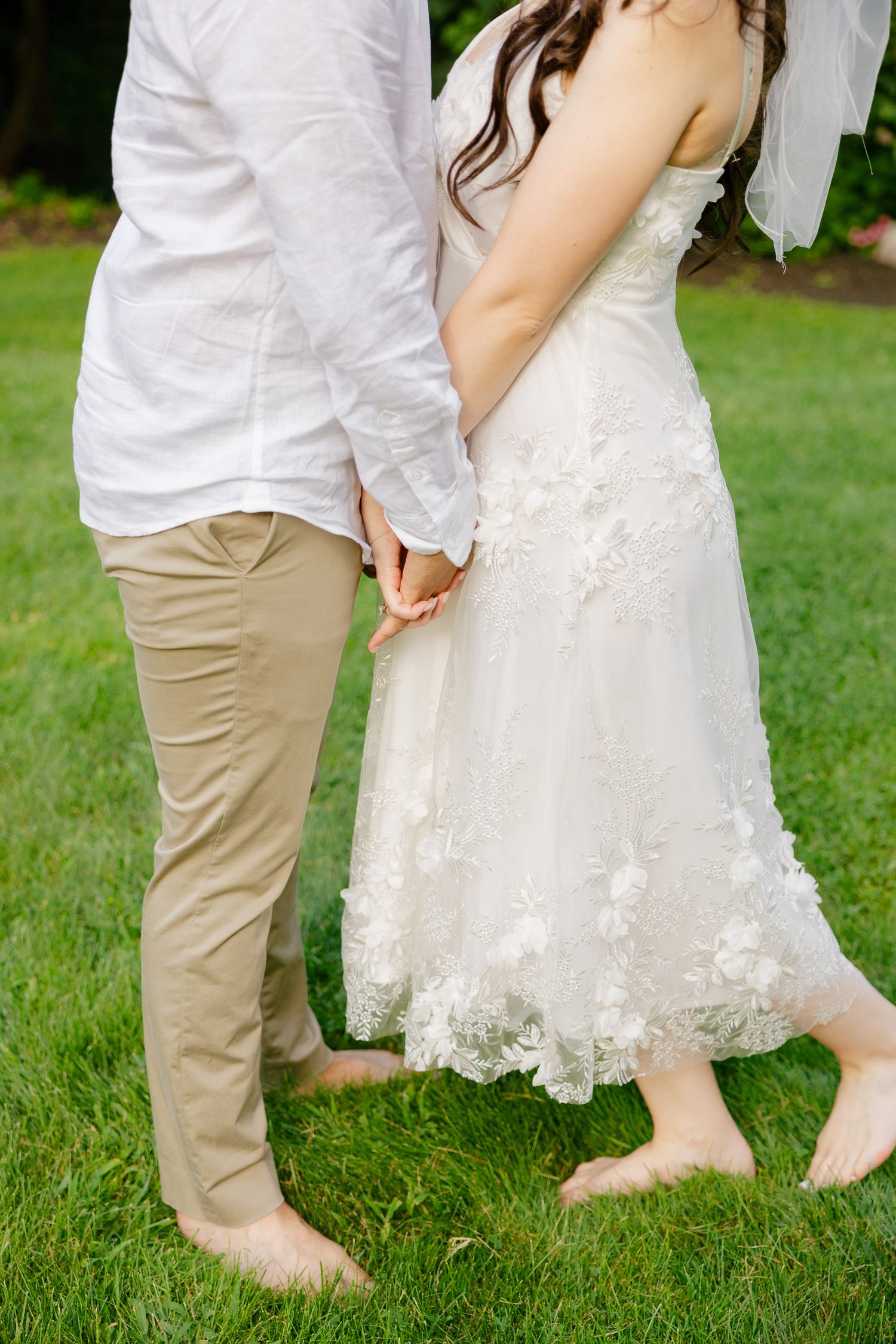 Close-up of couple holding hands outdoors, showing details of lace wedding dress and casual groomswear.