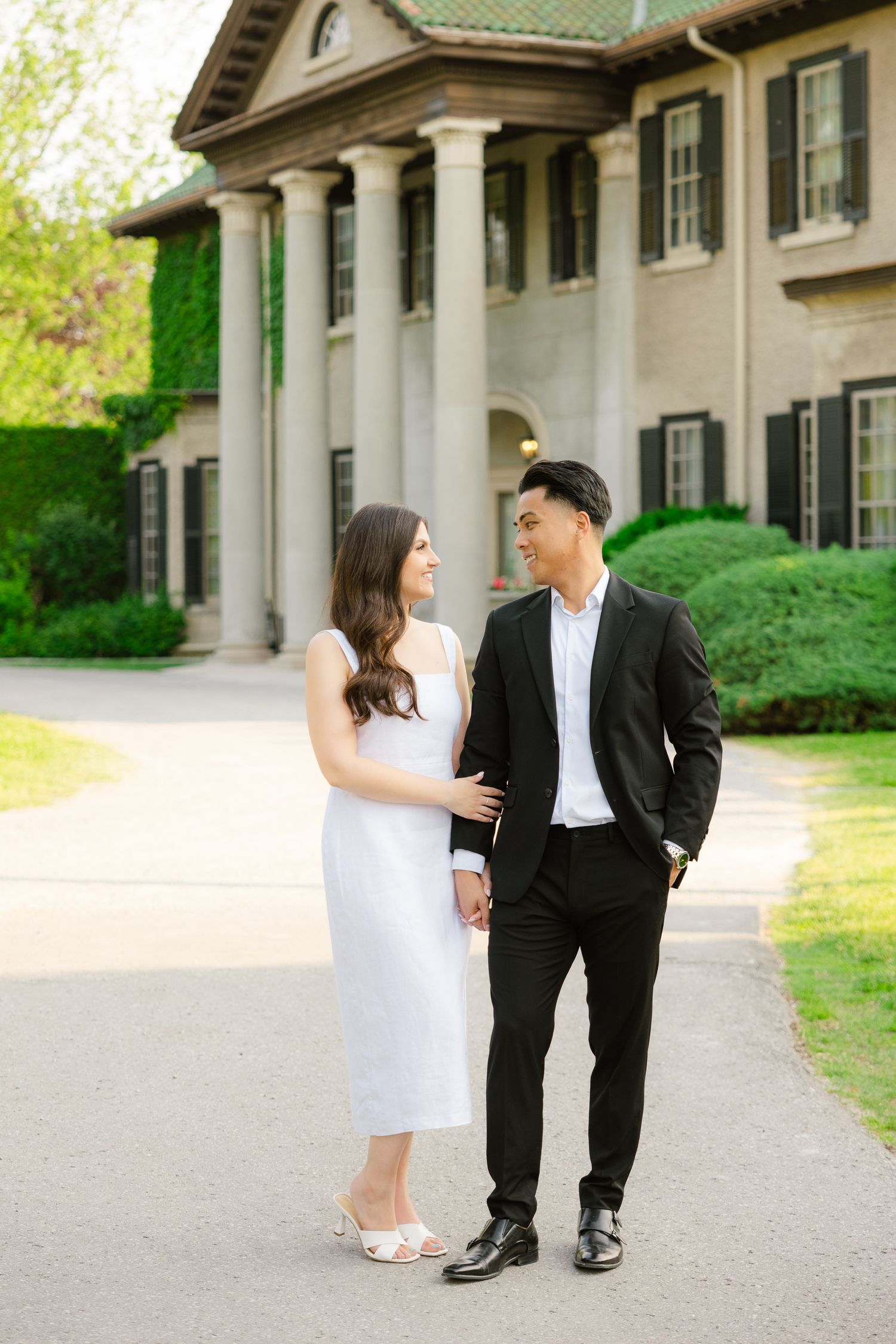 Couple shares a laugh while walking along pathway in front of stately mansion.